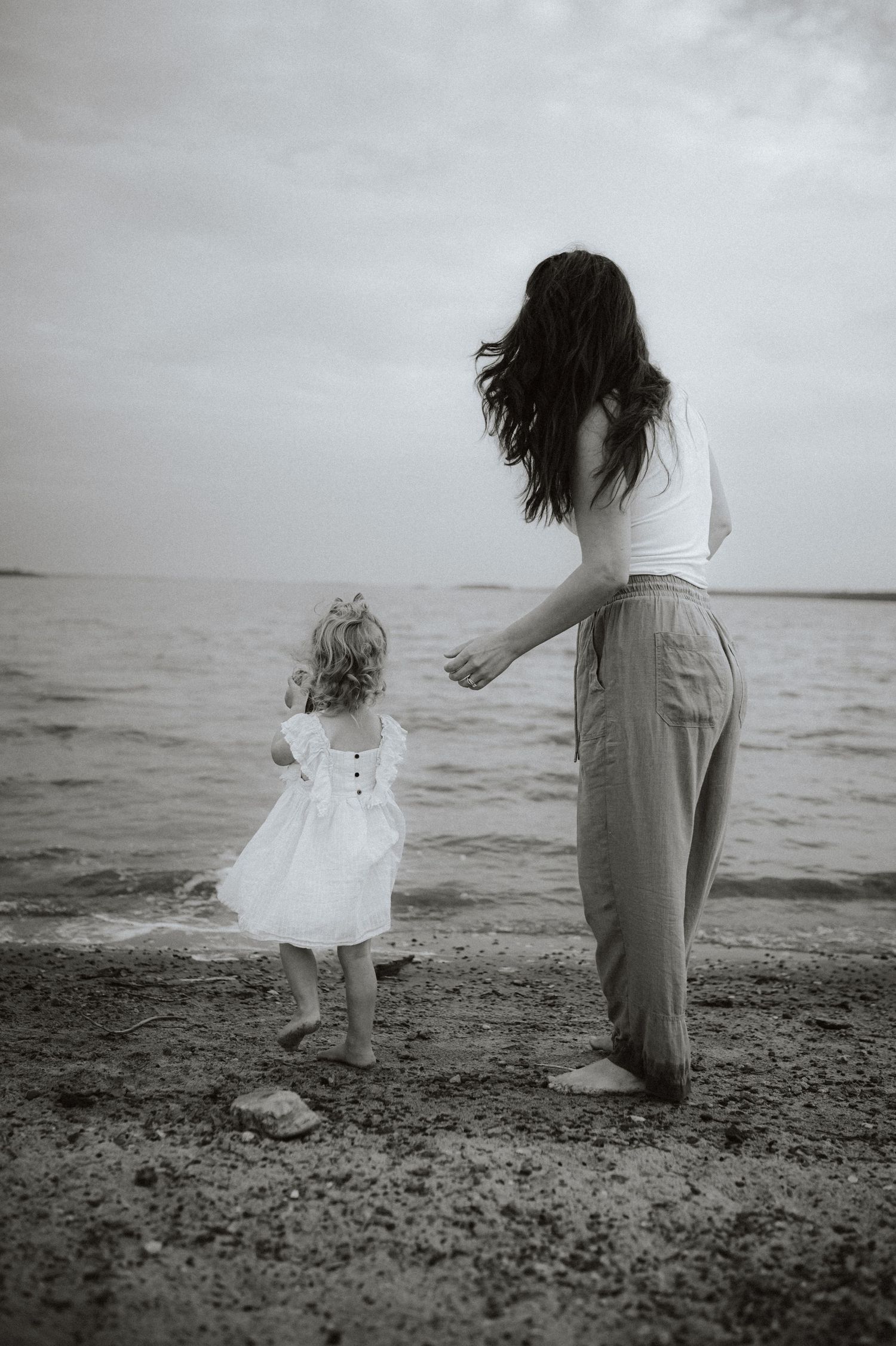 A woman and small child in white dress stand together on a rocky beach shore looking out at the ocean in black and white.