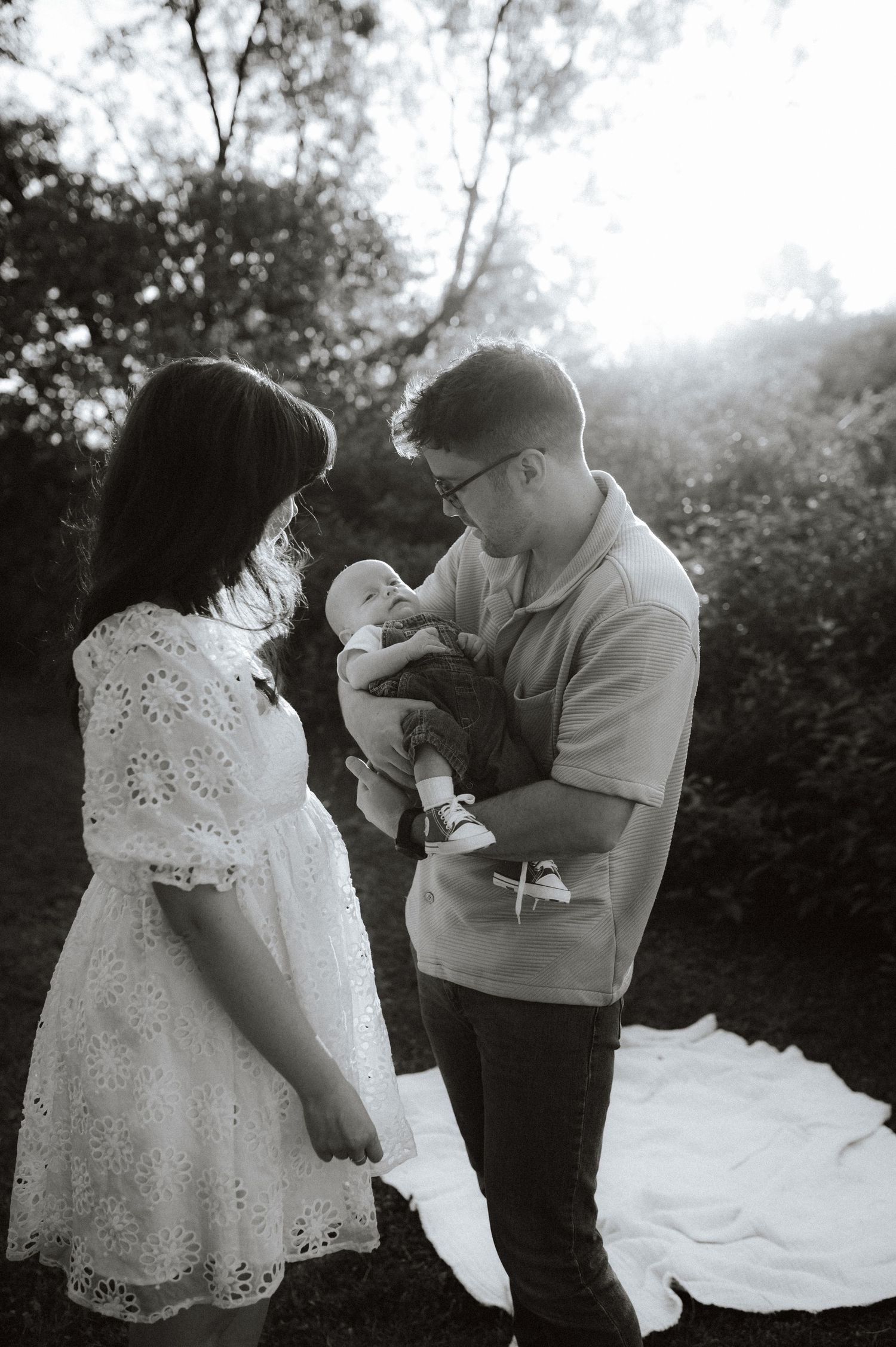 Black and white outdoor family portrait with a couple tenderly holding their infant on a blanket in a garden setting.