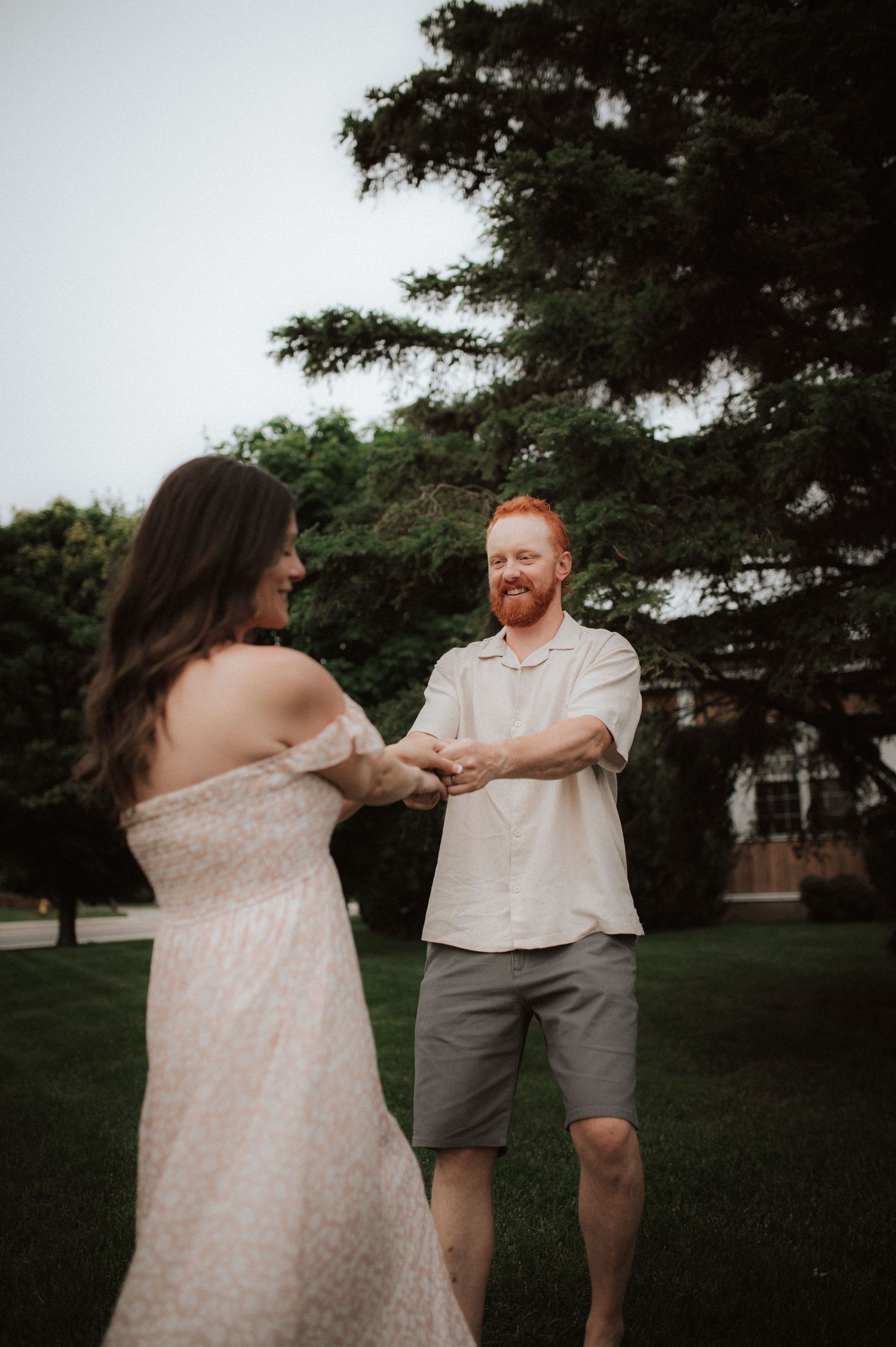 A couple in light colored outfits dances playfully together in a park setting at dusk.
