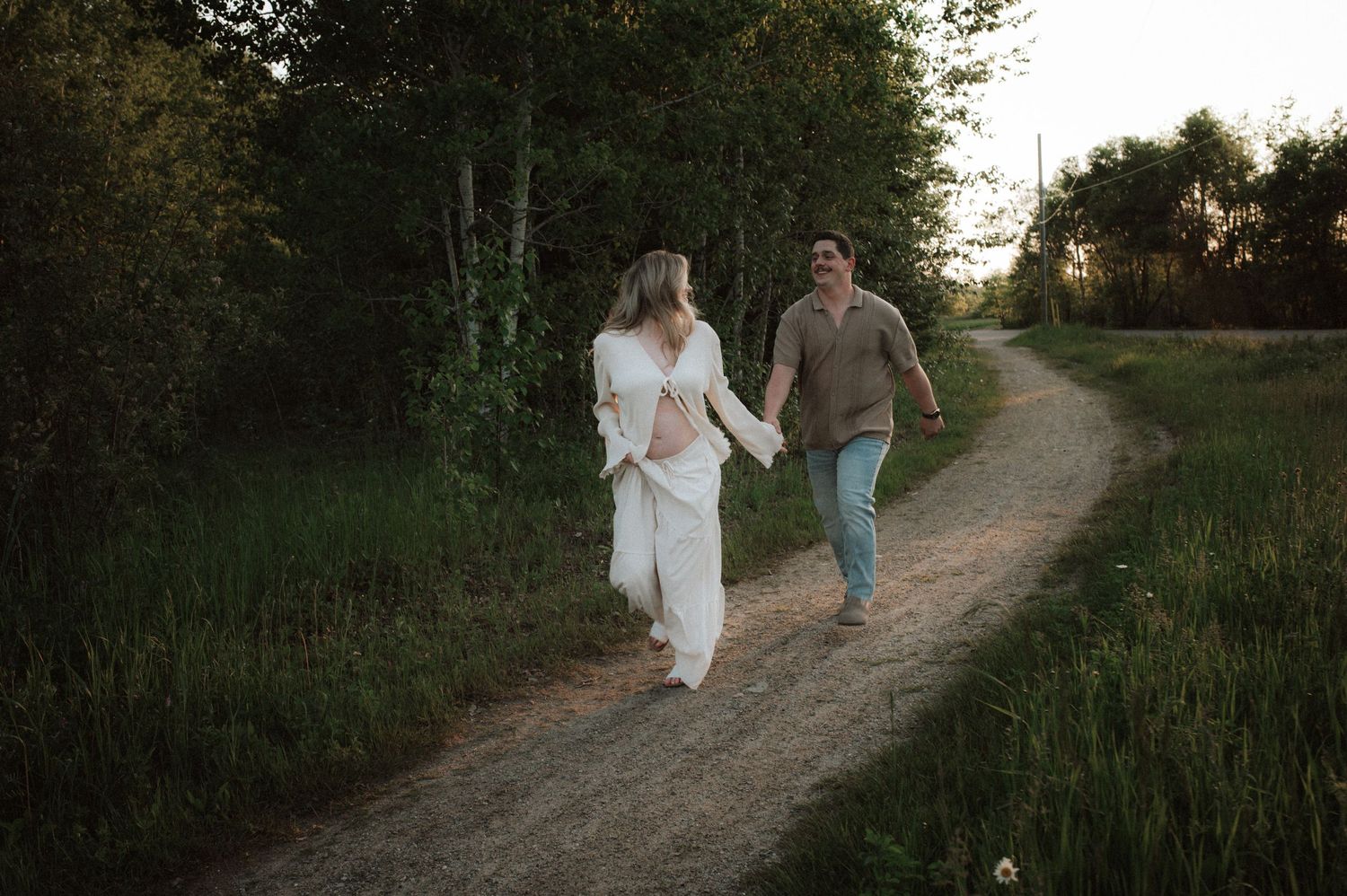 Two people in light-colored clothing holding hands while walking together on a dirt path at sunset in a natural setting.
