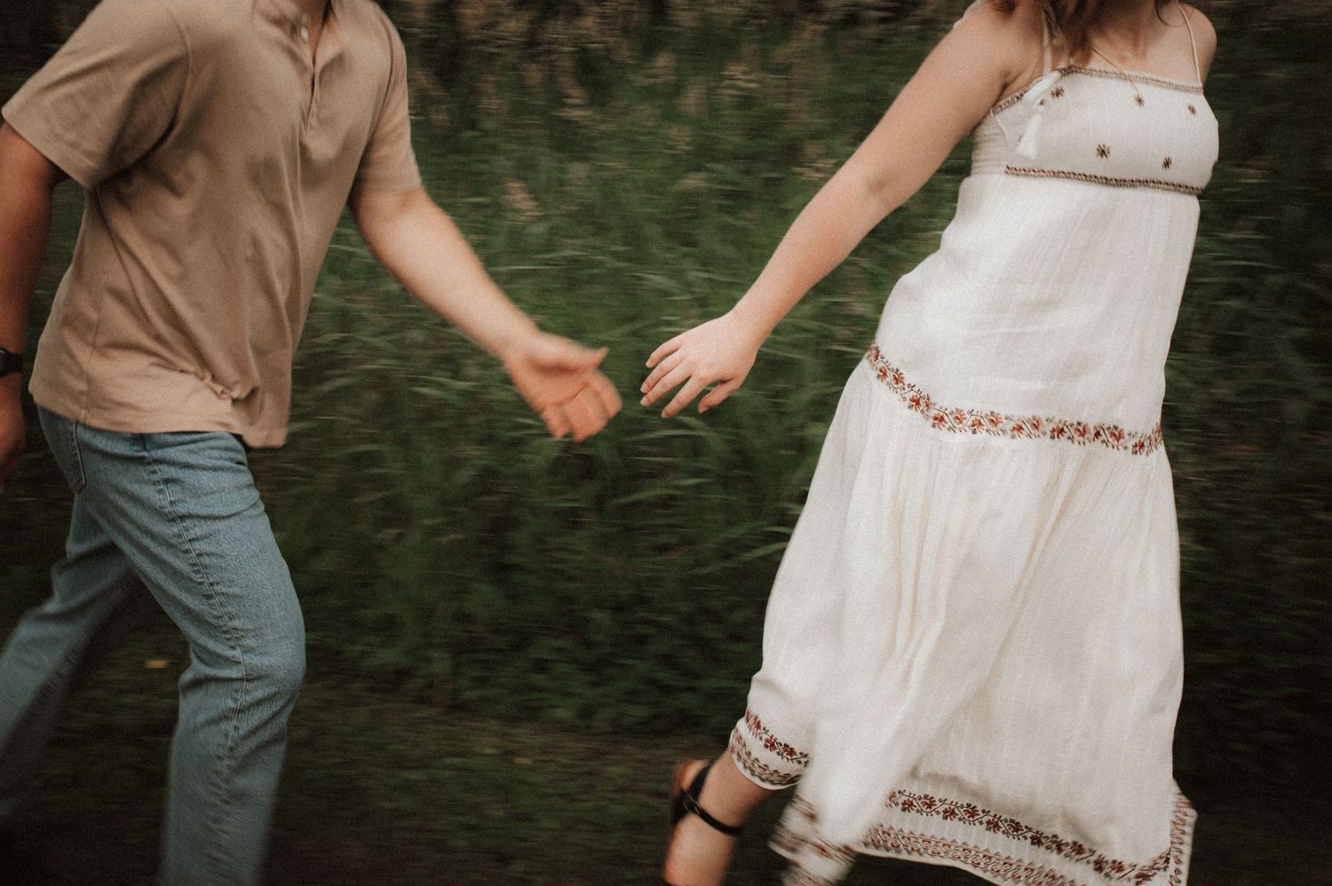 A couple holds hands while walking through grass, with one wearing a white summer dress and the other in casual attire.