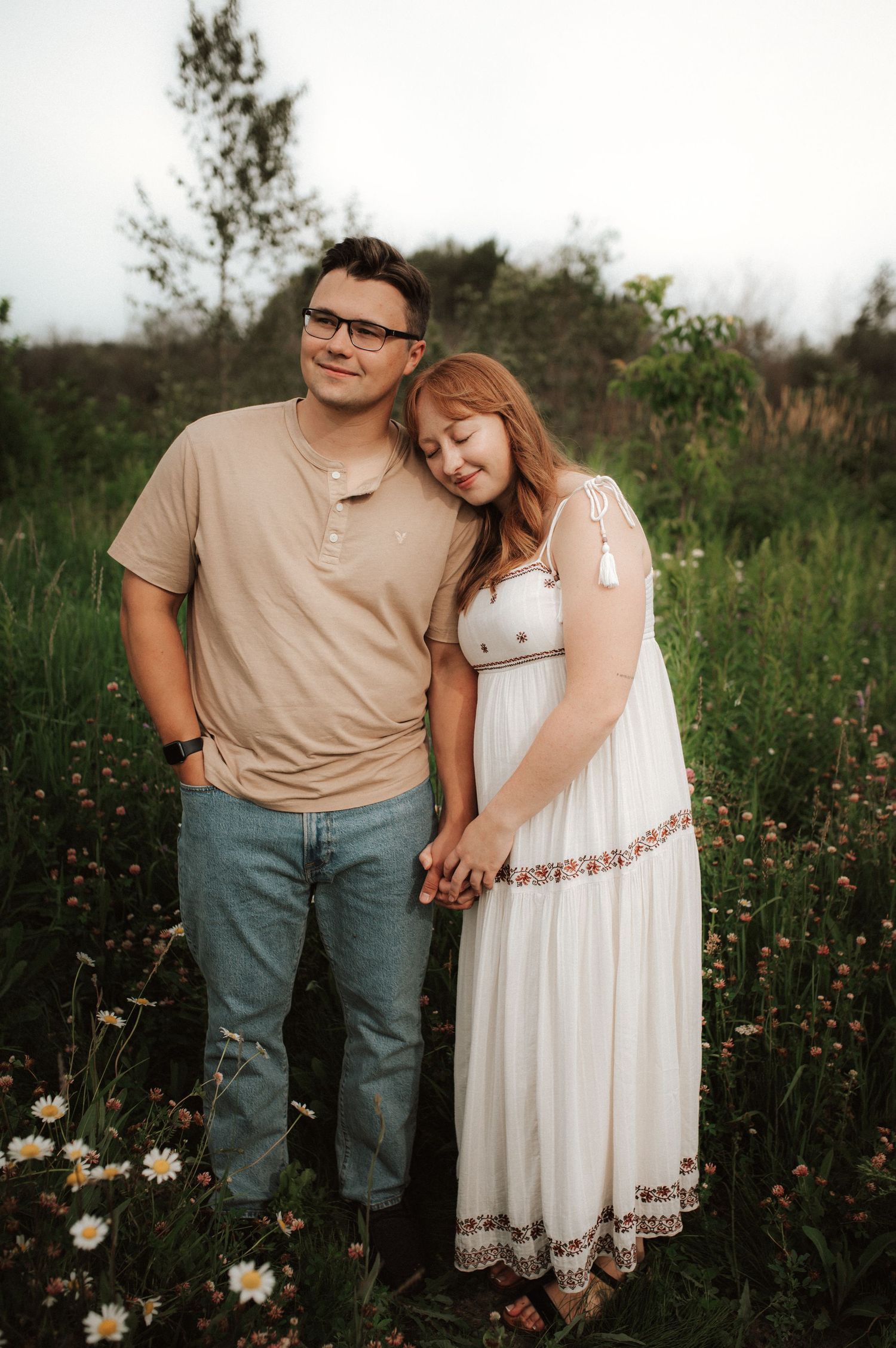 A romantic couple stands together in a wildflower field at sunset, wearing casual clothing and embracing tenderly.
