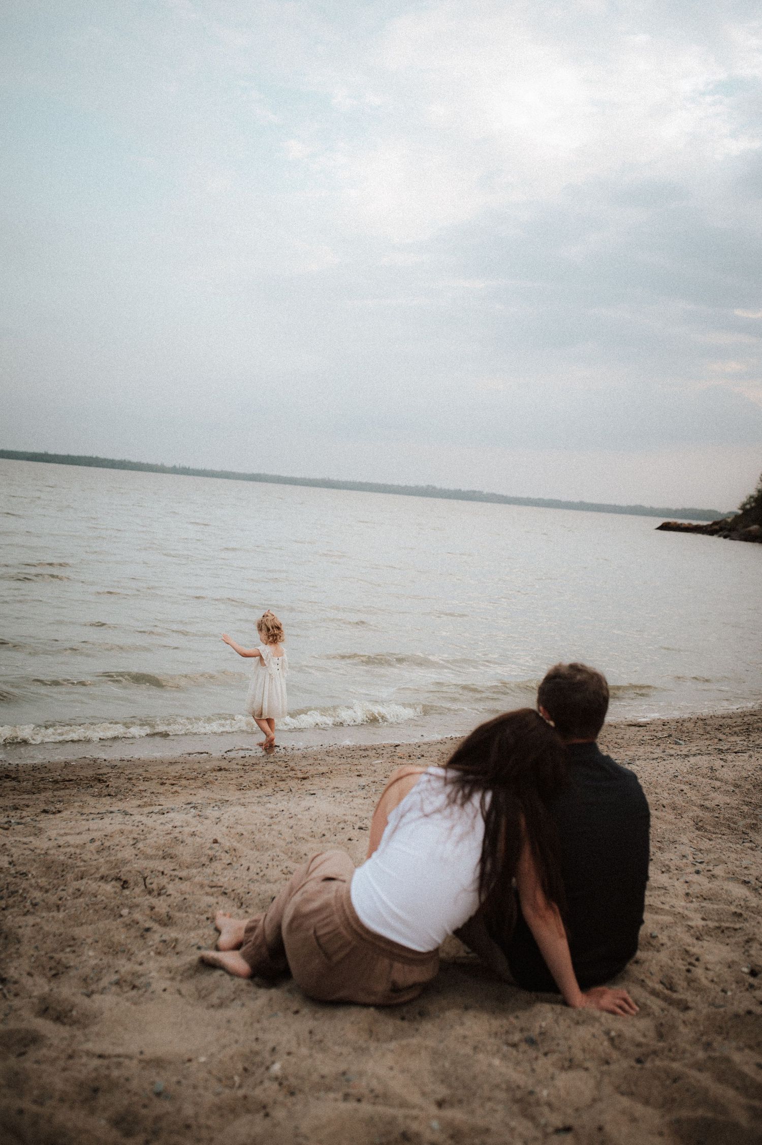 A couple sits on a sandy beach watching a small figure play near the water's edge during a cloudy day.