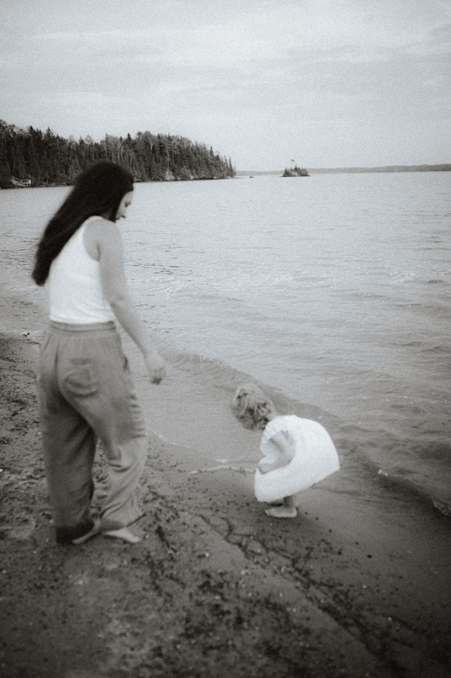 A black and white photo shows two figures on a rocky beach shore overlooking a serene lake with forested coastline.