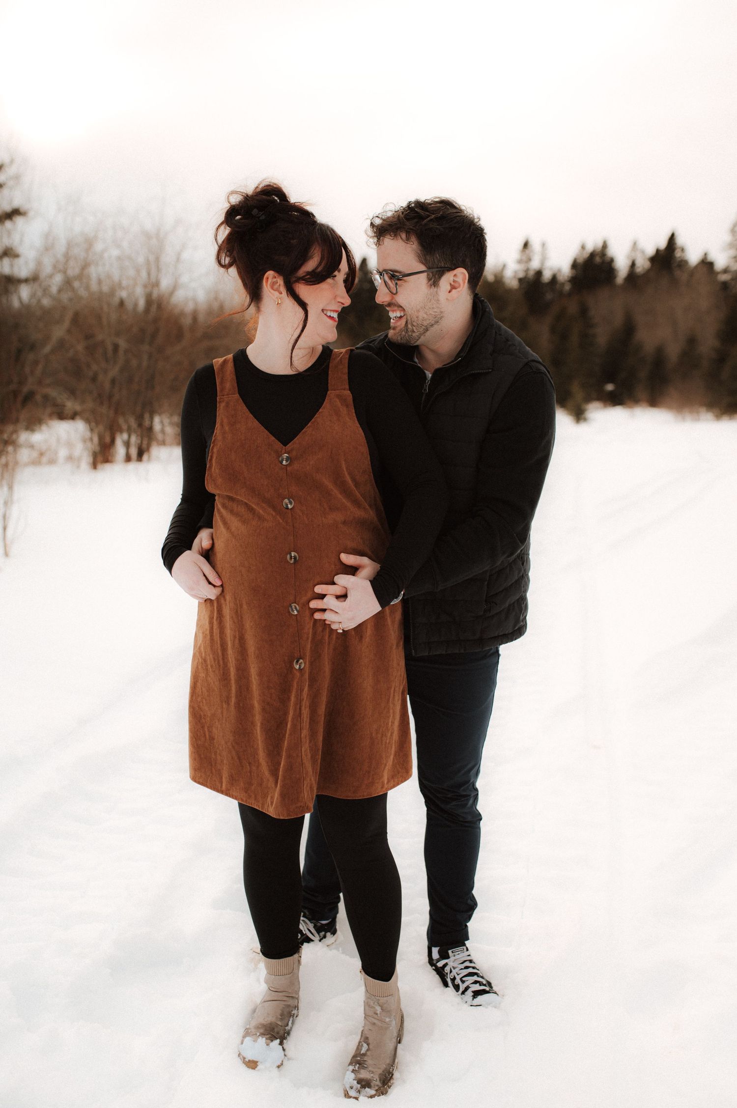 A couple share an intimate winter moment outdoors in the snow, wearing cozy layers and a brown corduroy dress.