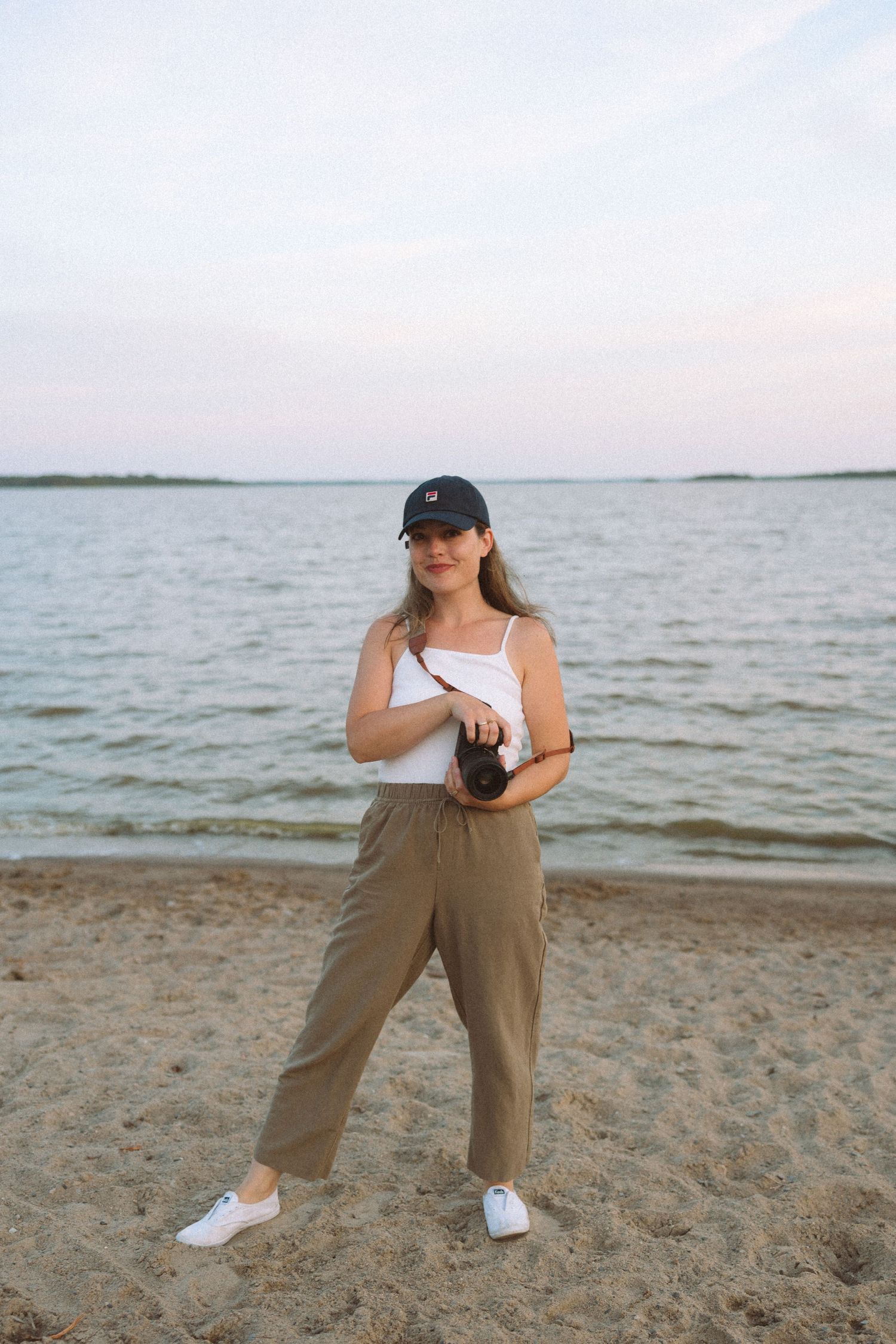 Person in casual attire with baseball cap poses on sandy beach at sunset with water in background.