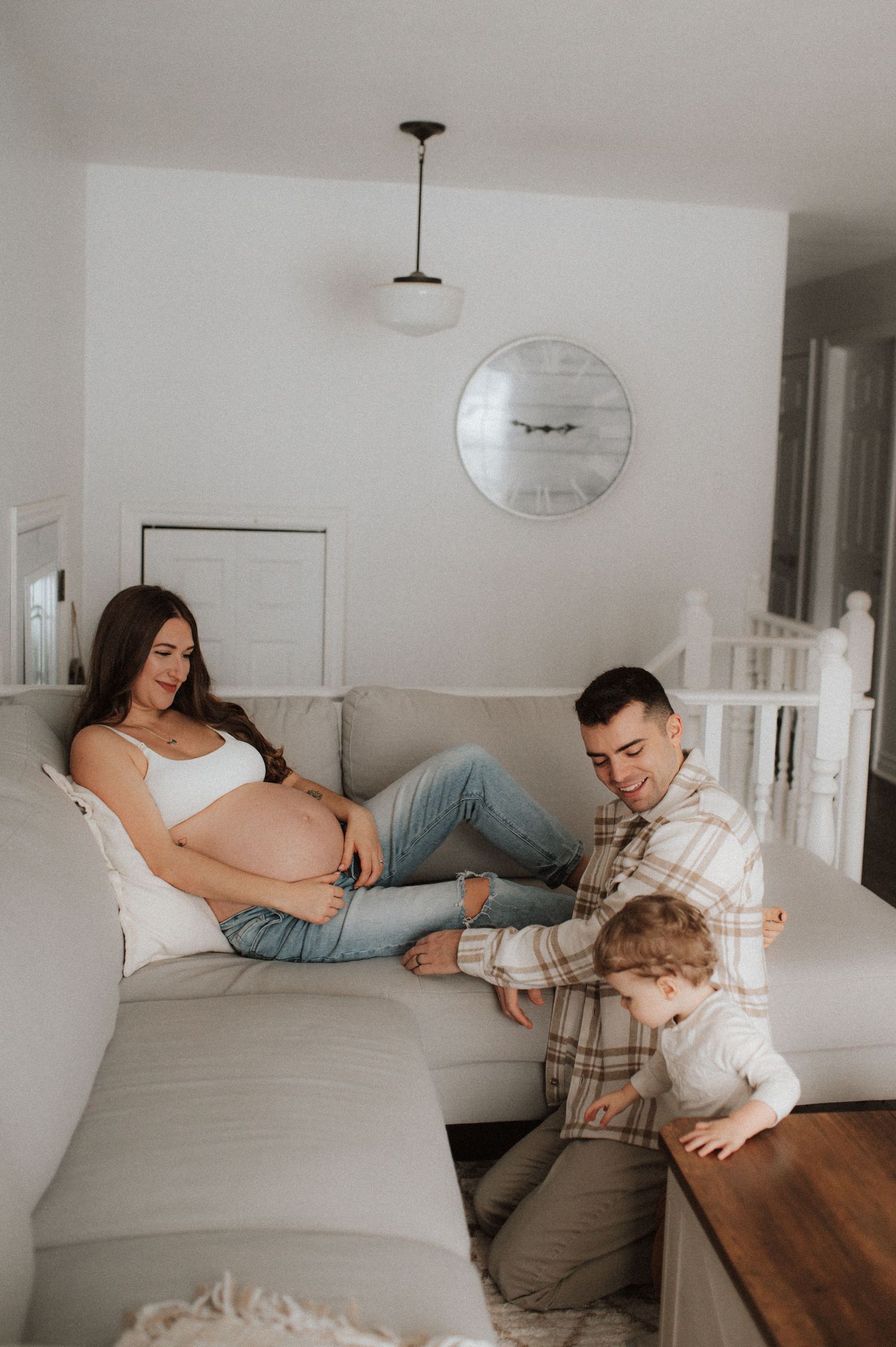 A pregnant woman relaxes on a gray sectional sofa at home while a family spends time together in the living room.