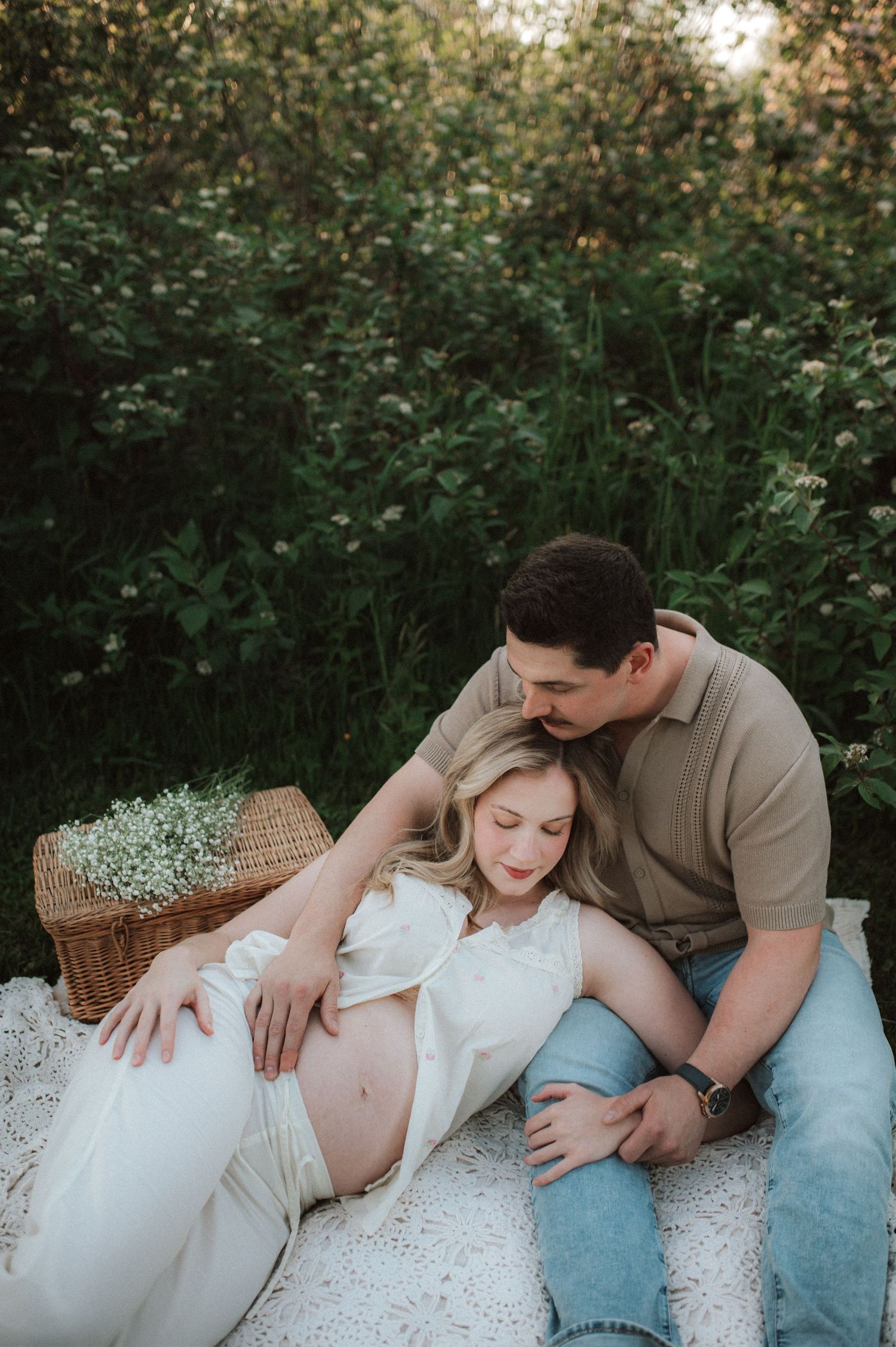 Expectant couple shares intimate moment during maternity photoshoot outdoors with picnic basket and white dress.