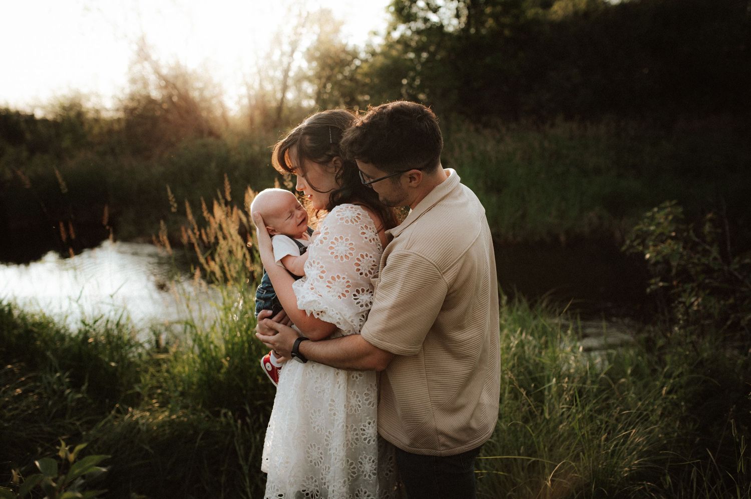 A romantic sunset moment with embracing figures in white lace dress and beige sweater beside a tranquil pond.