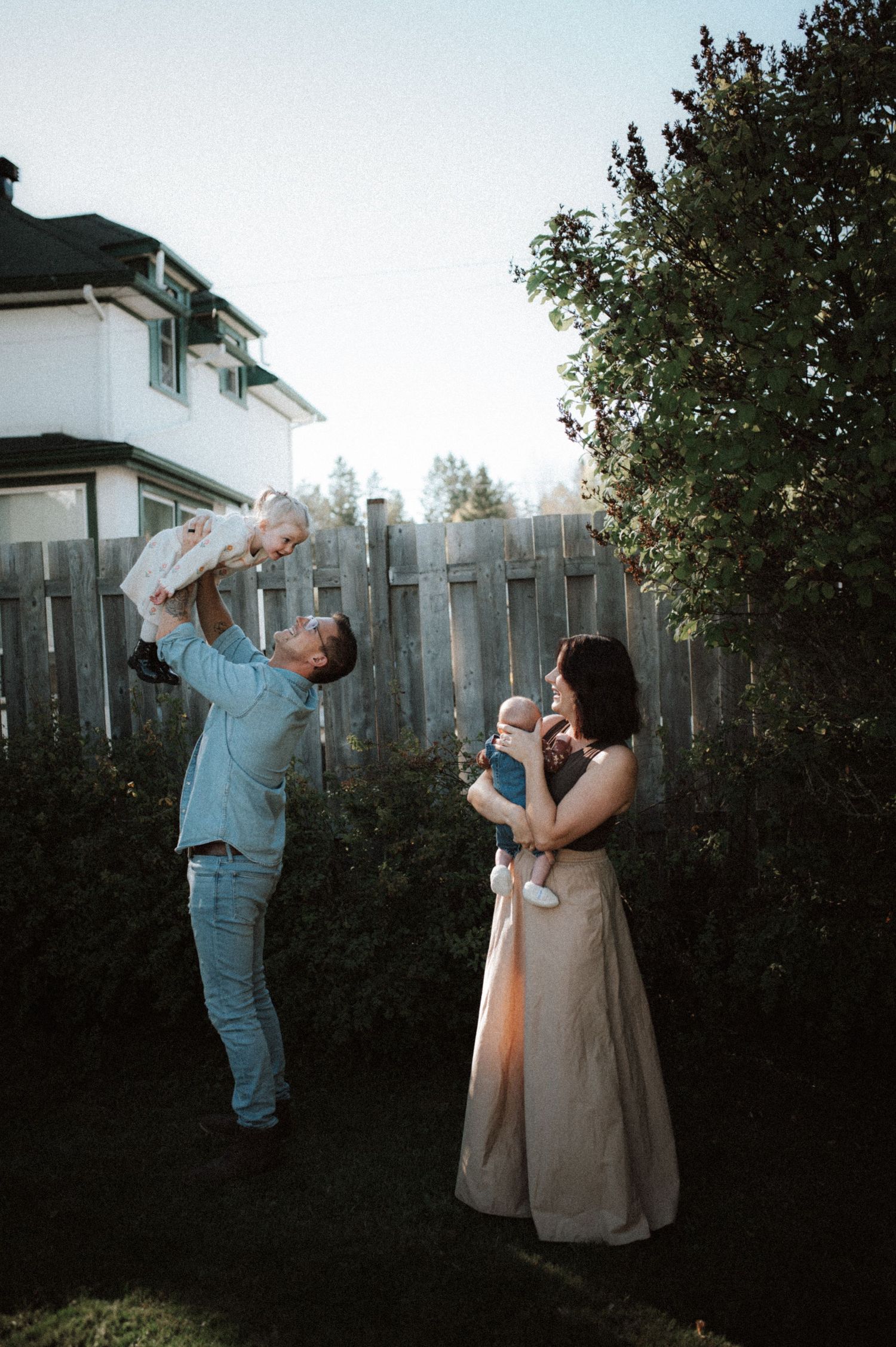 A couple stands in a backyard at sunset with babies, lifting them up and embracing them lovingly.
