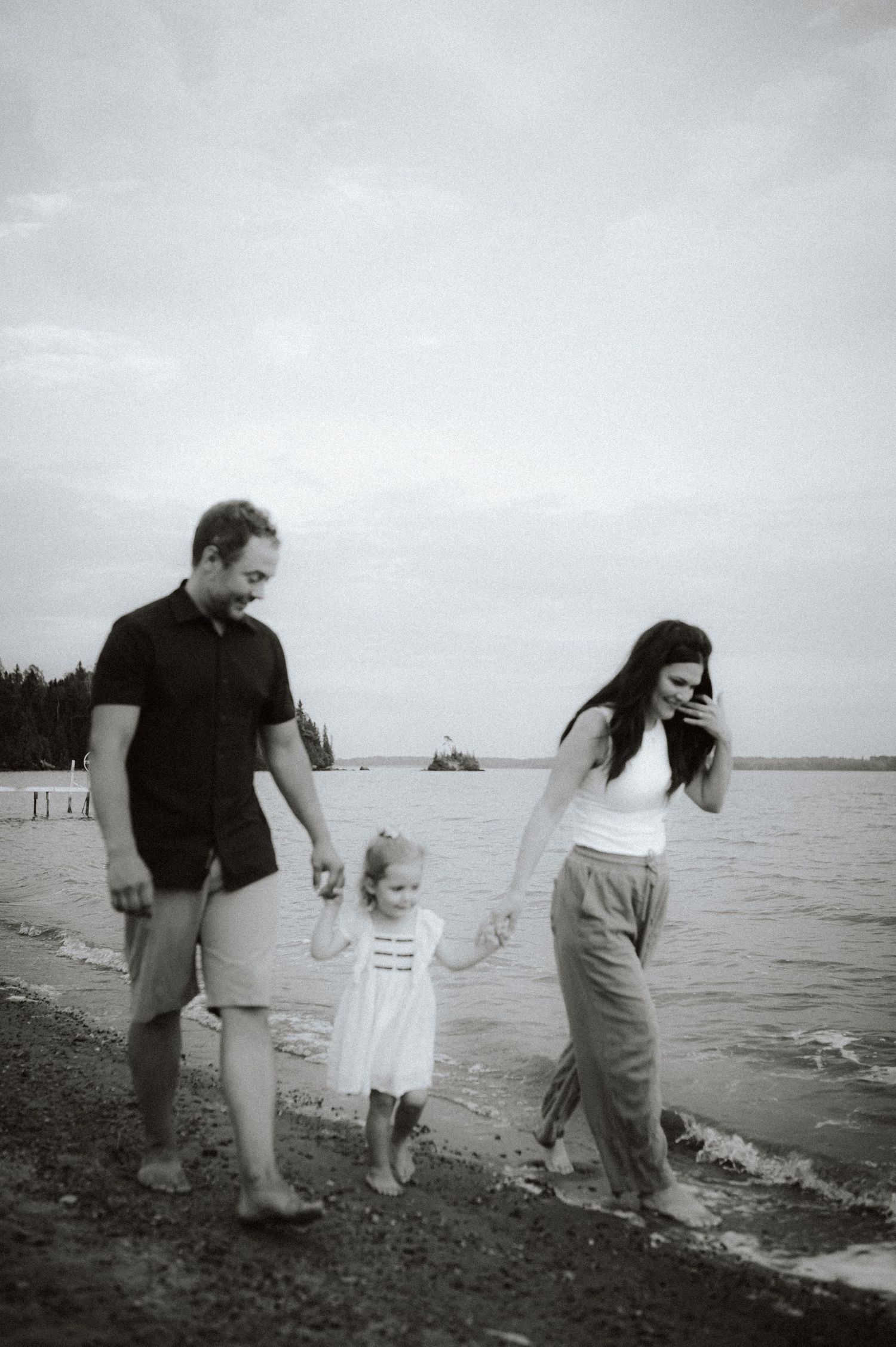 Family of three walk together holding hands along a moody beach shoreline in black and white.