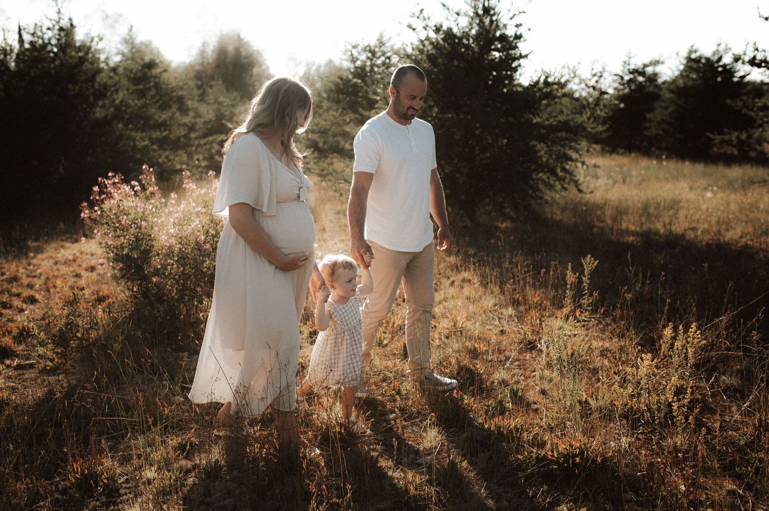 A family stands together in a sunlit field at golden hour with tall grass and trees in the background.