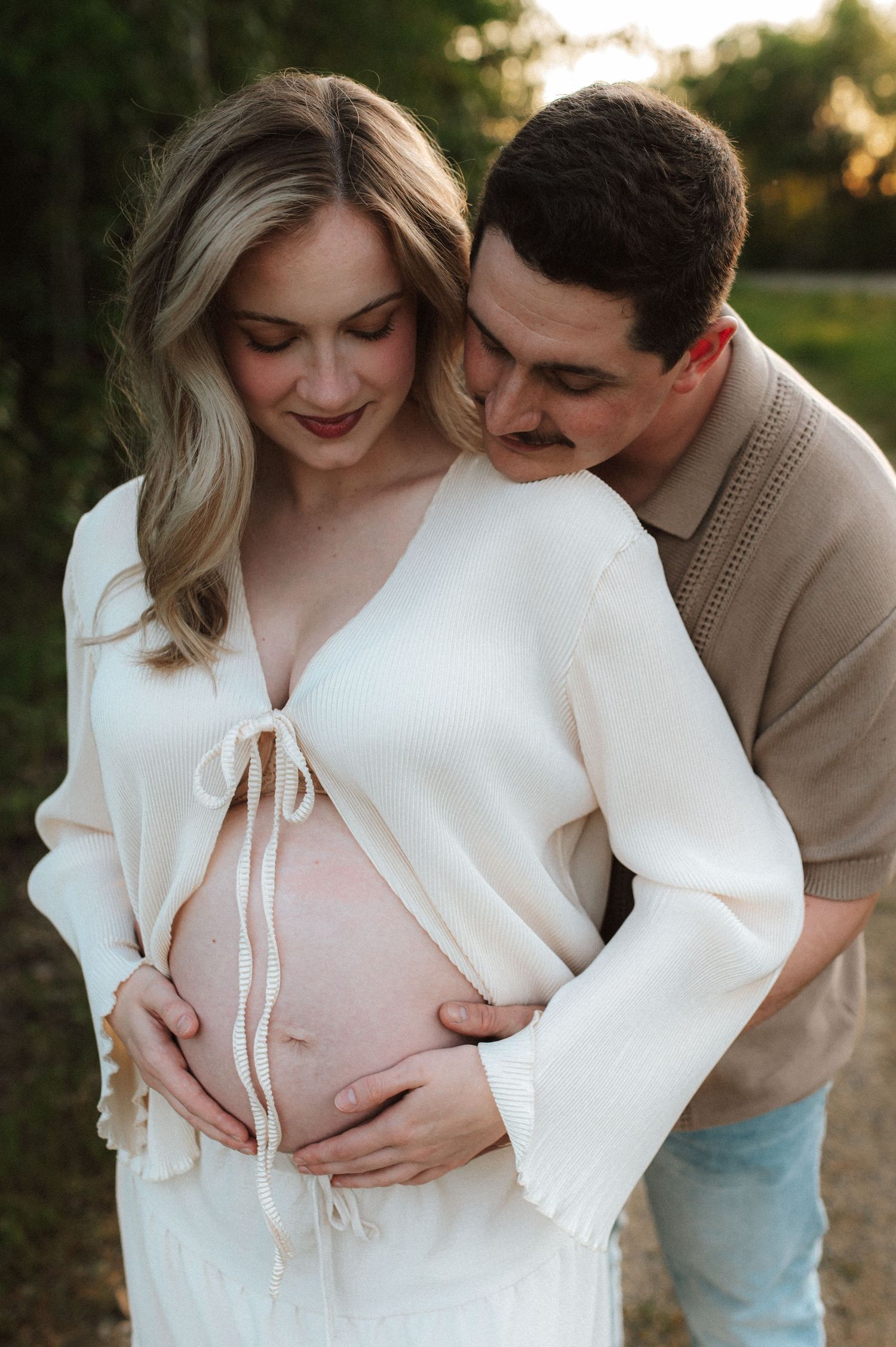 Couple in warm sunset light share intimate maternity photos outdoors wearing cream and beige attire.