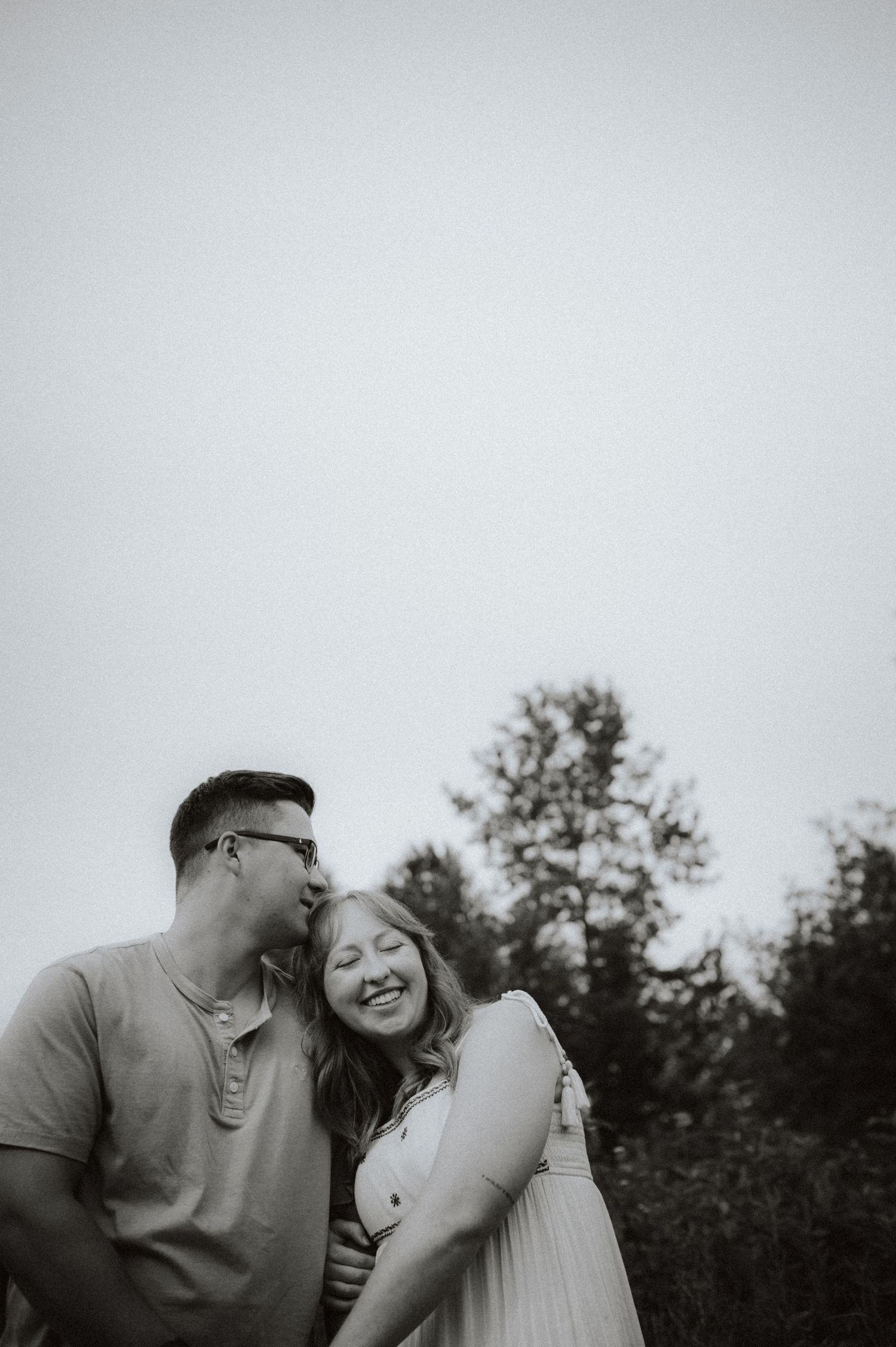 A tender black and white romantic moment captured outdoors with trees in the background.