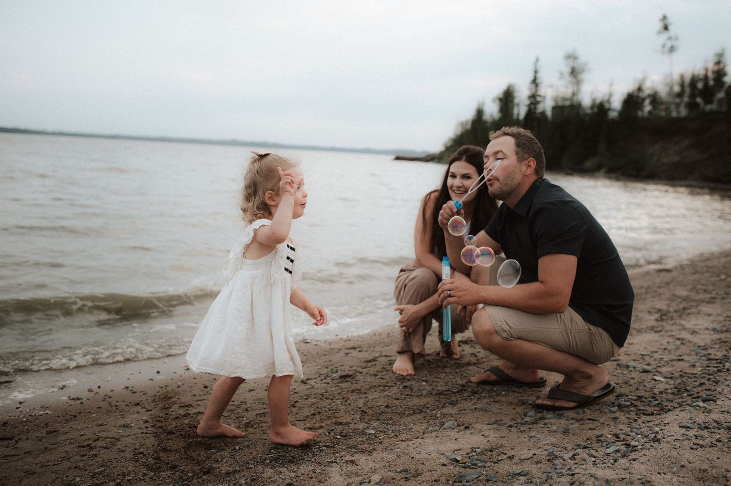 A toddler in a white dress plays on a beach at sunset while a couple crouches nearby on the sandy shoreline.