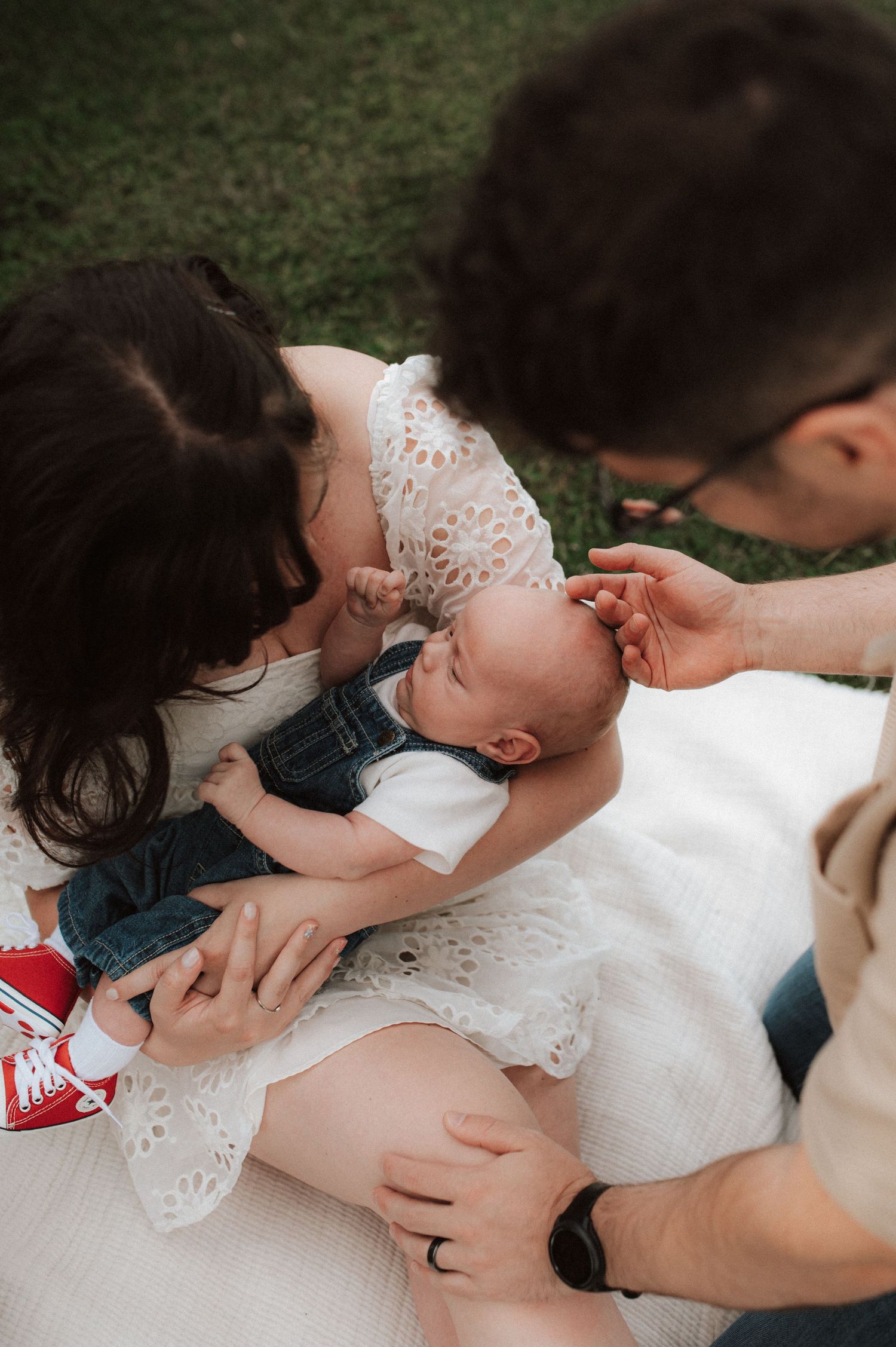 A tender family moment on a blanket outdoors showing parents lovingly embracing their infant in white clothing.