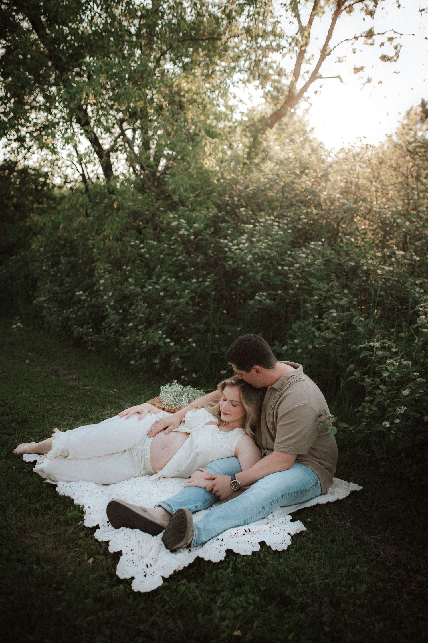 A couple relaxes on a white blanket in a garden setting during golden hour sunlight filtering through trees.