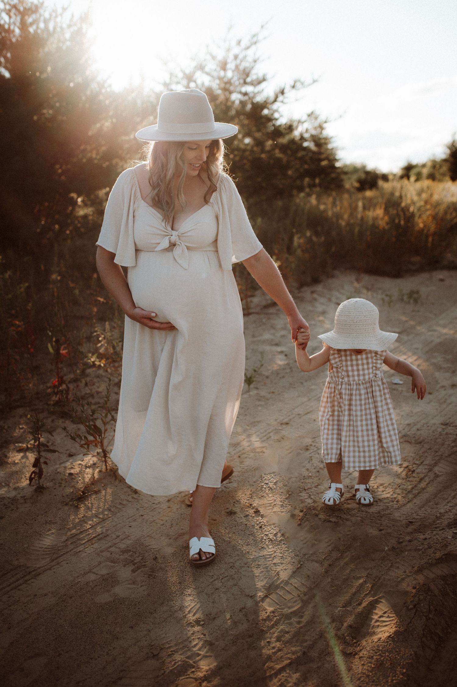 A figure in white clothing walks hand in hand with a small child on a sunlit dirt path at sunset.
