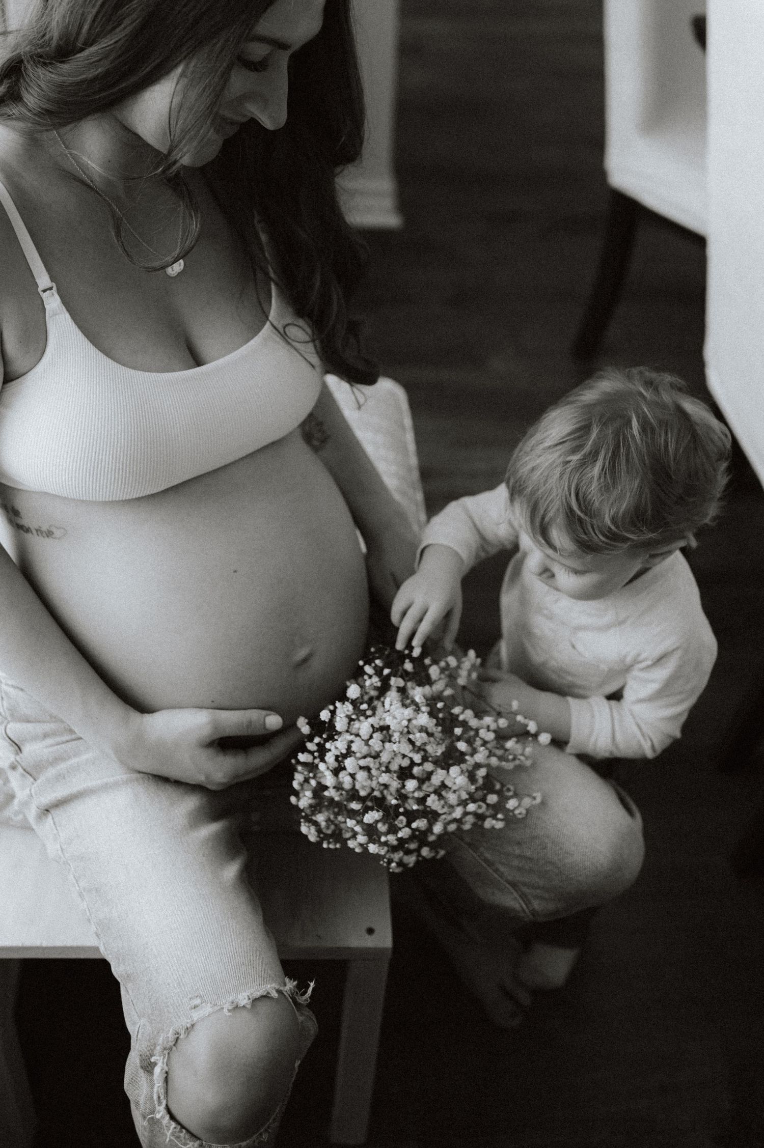 Black and white photo of a pregnant woman in a white top holding flowers with a small child touching her belly.
