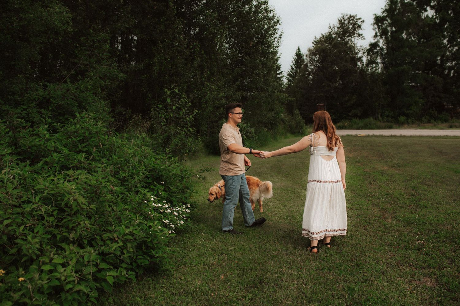 Two people hold hands in a romantic moment on a grassy path surrounded by lush greenery and trees.