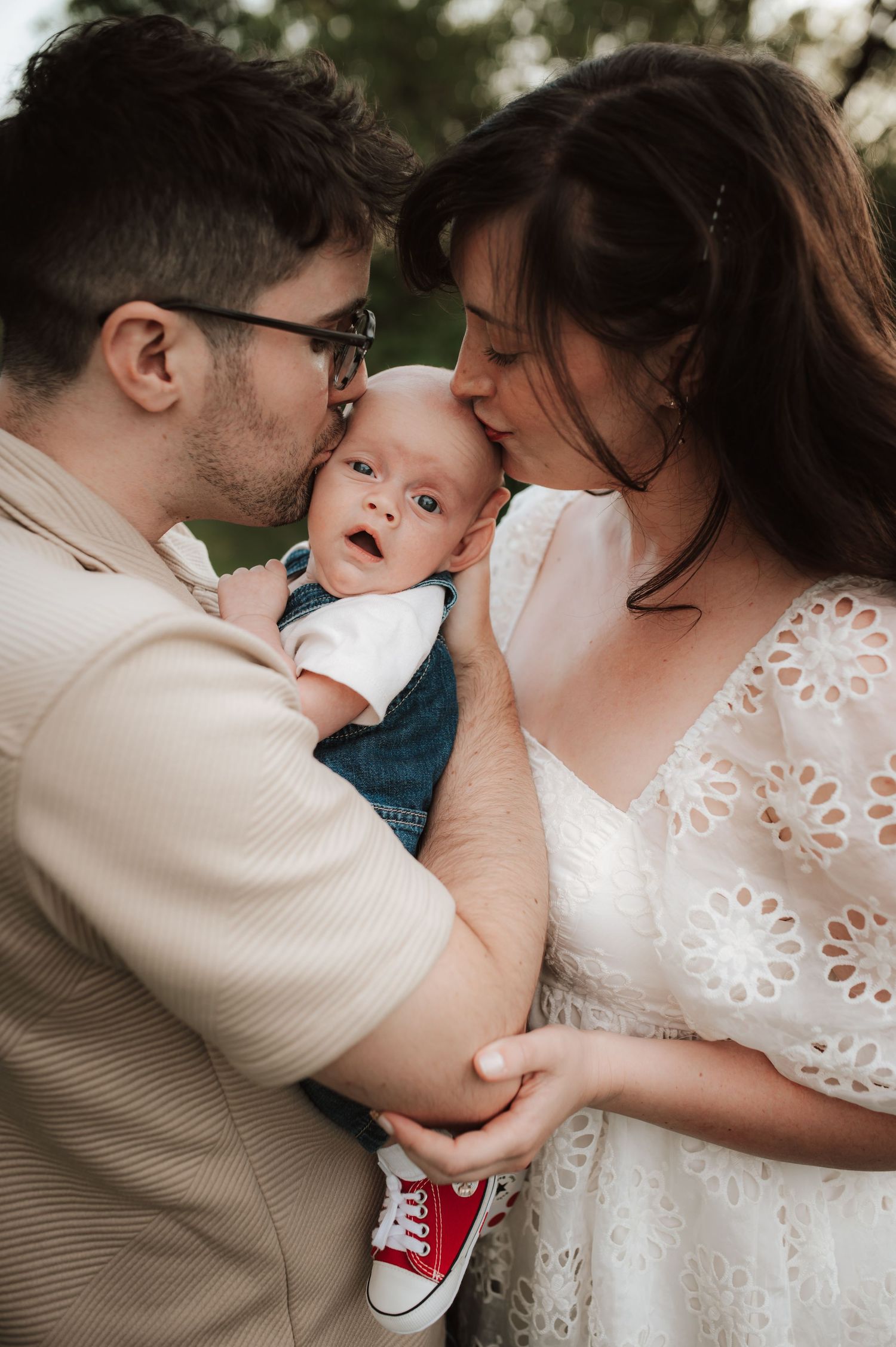 A sweet family moment as parents embrace their baby wearing red sneakers and a denim outfit in a romantic outdoor setting.