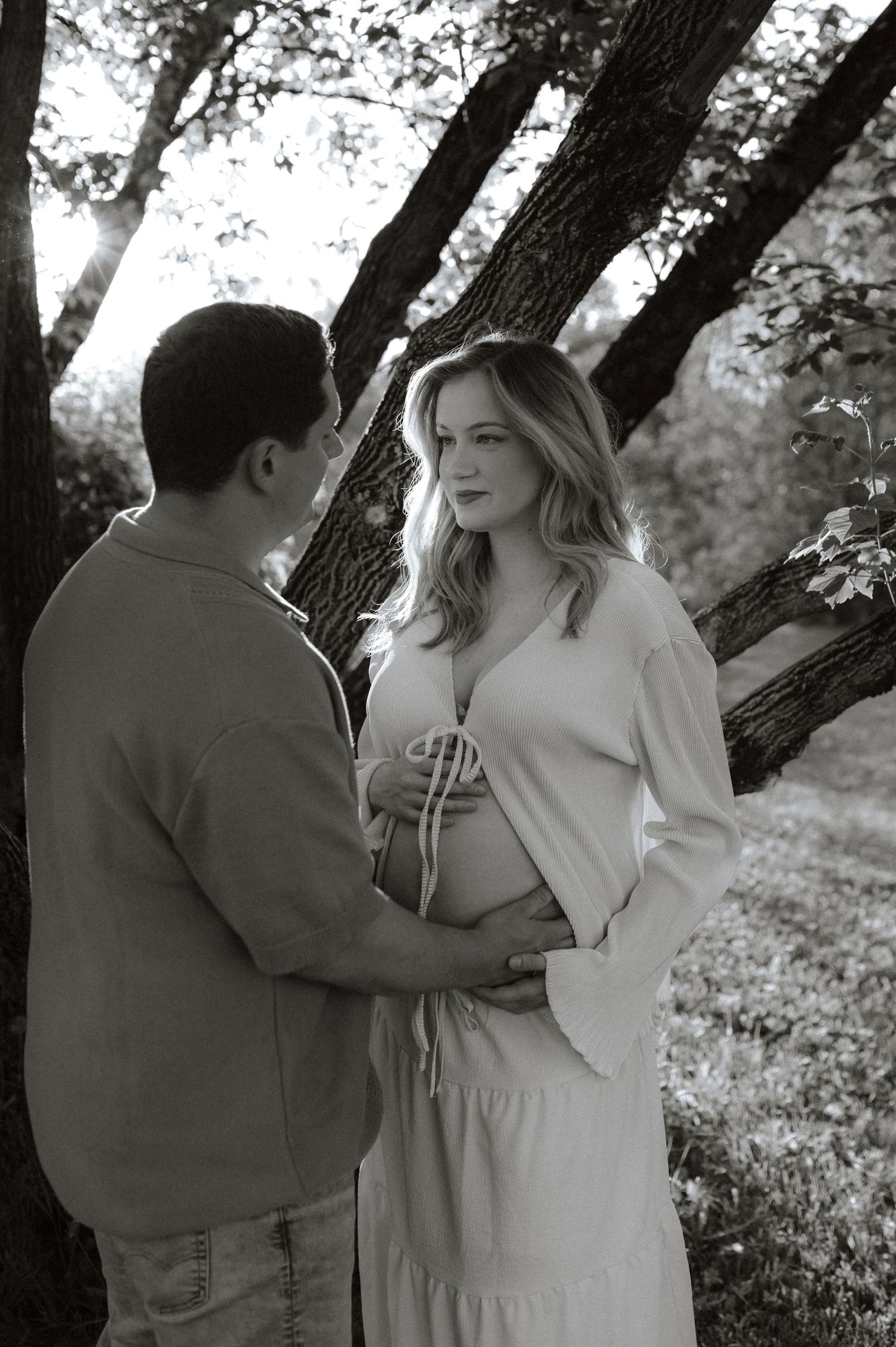 A black and white maternity photo with a couple standing near a tree as sunlight filters through the branches.