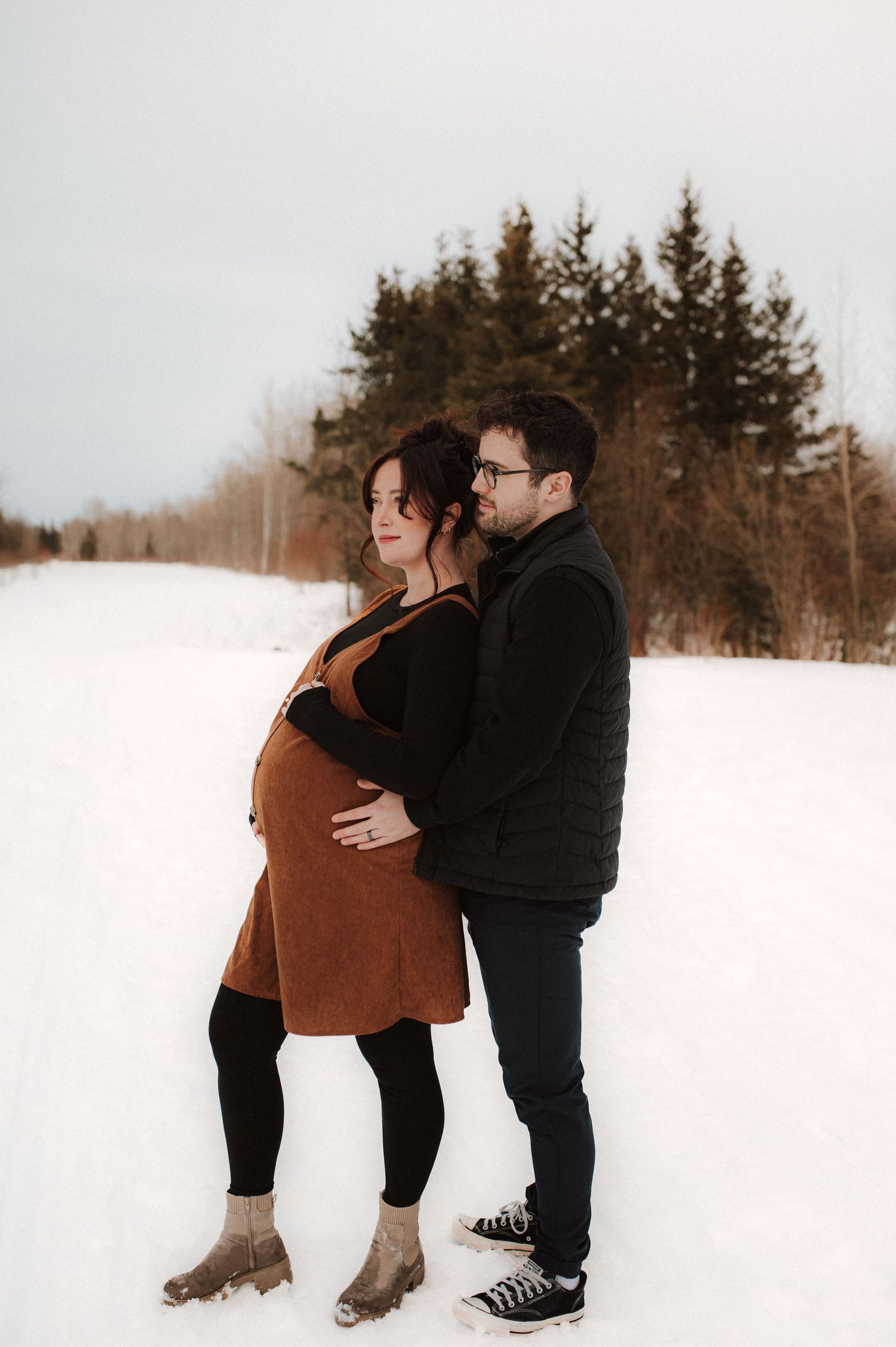 A couple embraces in a snowy winter landscape while taking maternity photos in the forest.