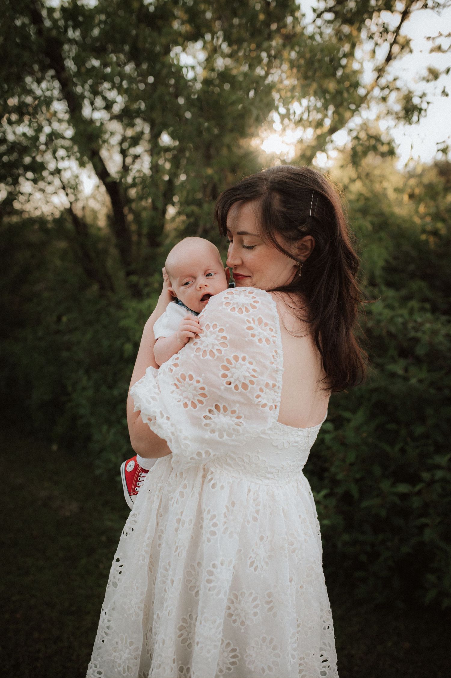 A tender moment is shared in white eyelet lace dresses against a natural outdoor backdrop at sunset.