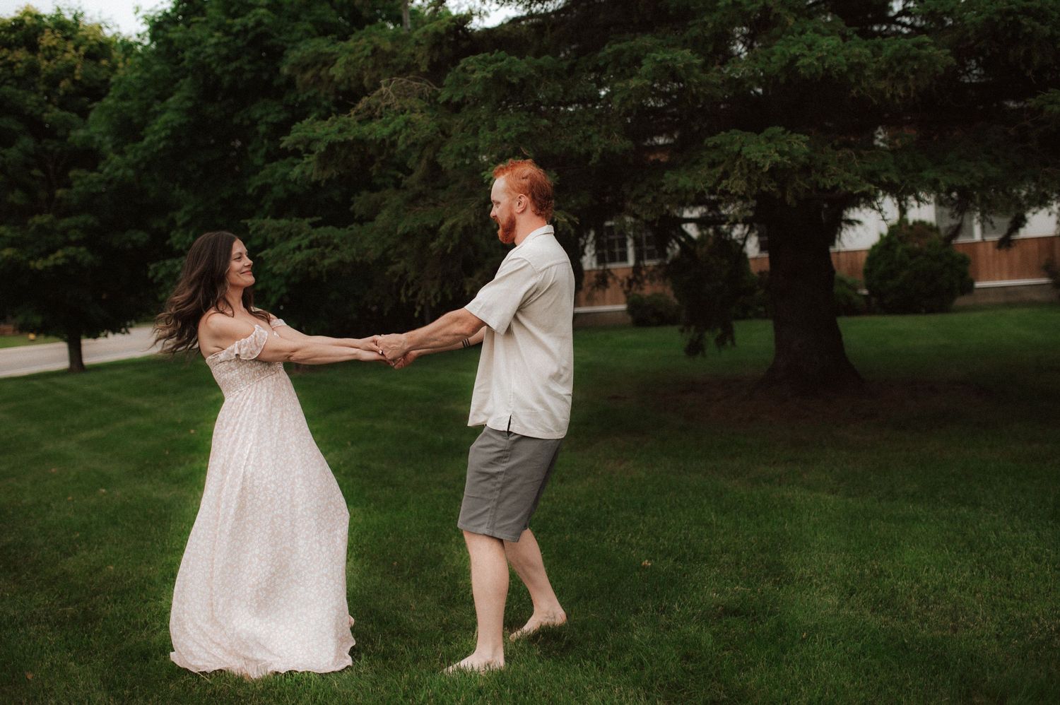 A couple holds hands while dancing together on a grass lawn surrounded by trees on a summer evening.