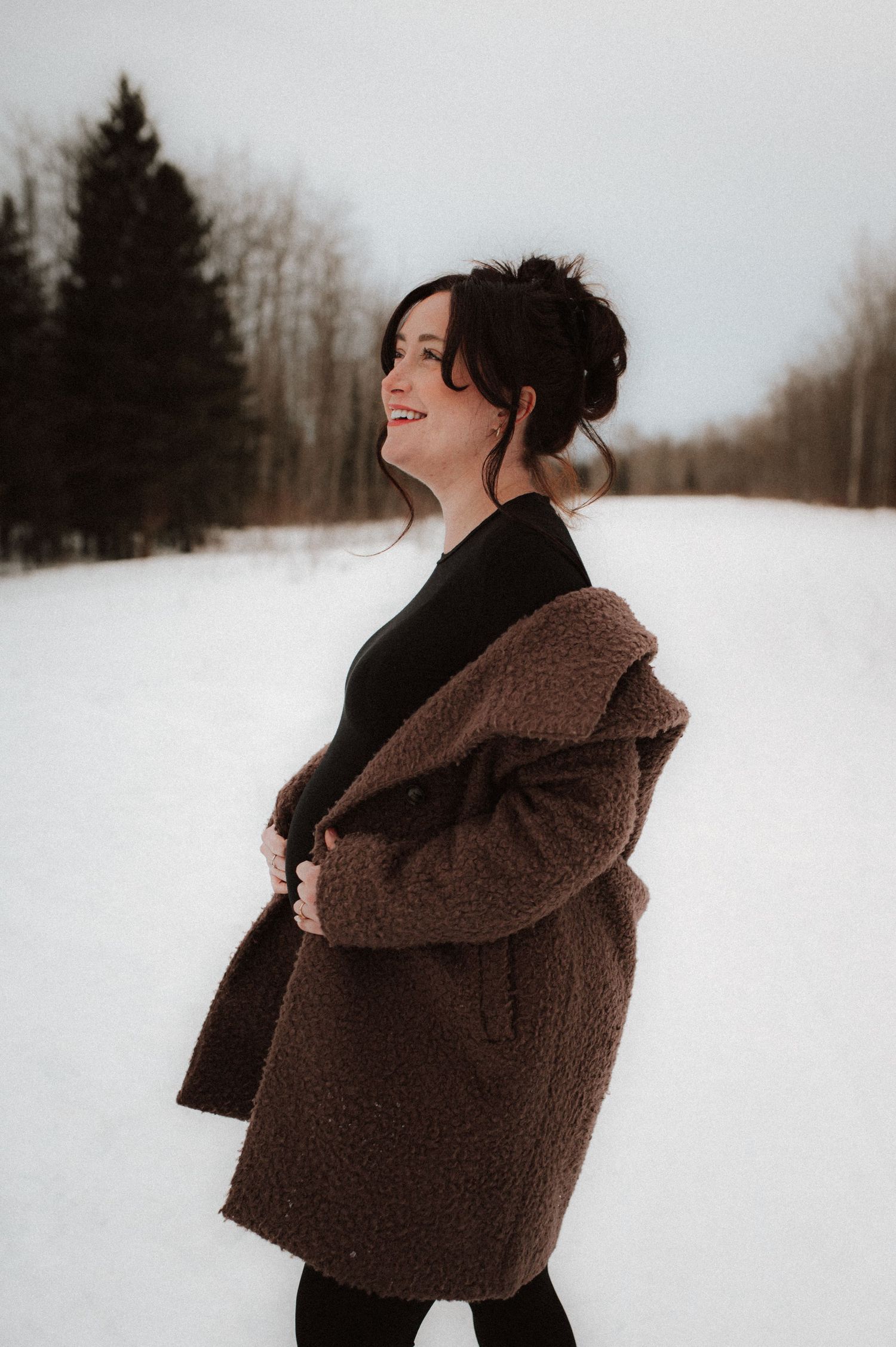 A person in a brown winter coat stands against a snowy winter landscape in moody black and white photography.