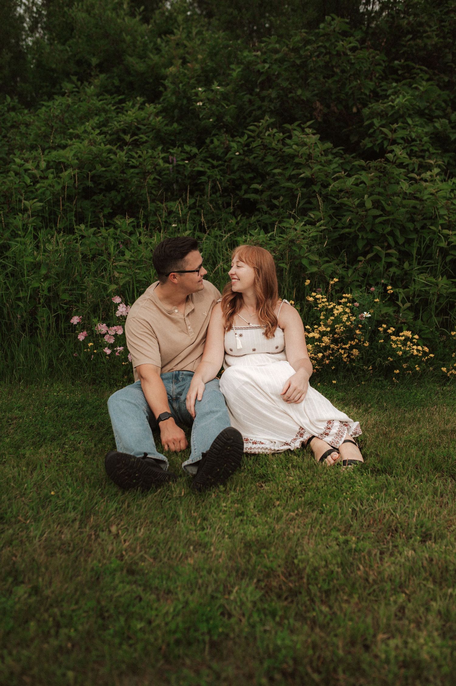 A couple sits together lovingly on grass with orange flowers blooming in the garden background.