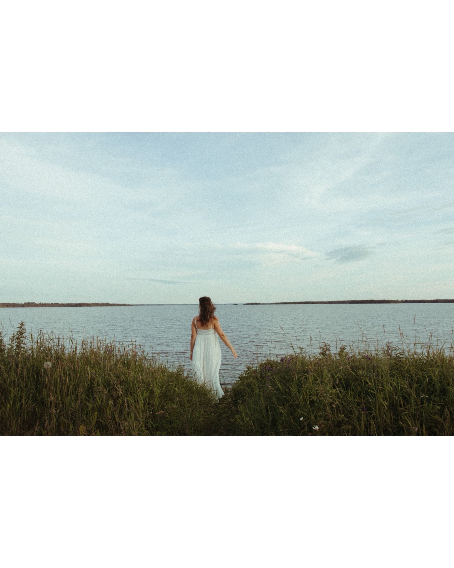 Person in a white dress stands along a serene coastline with tall grasses and calm waters stretching to the horizon.