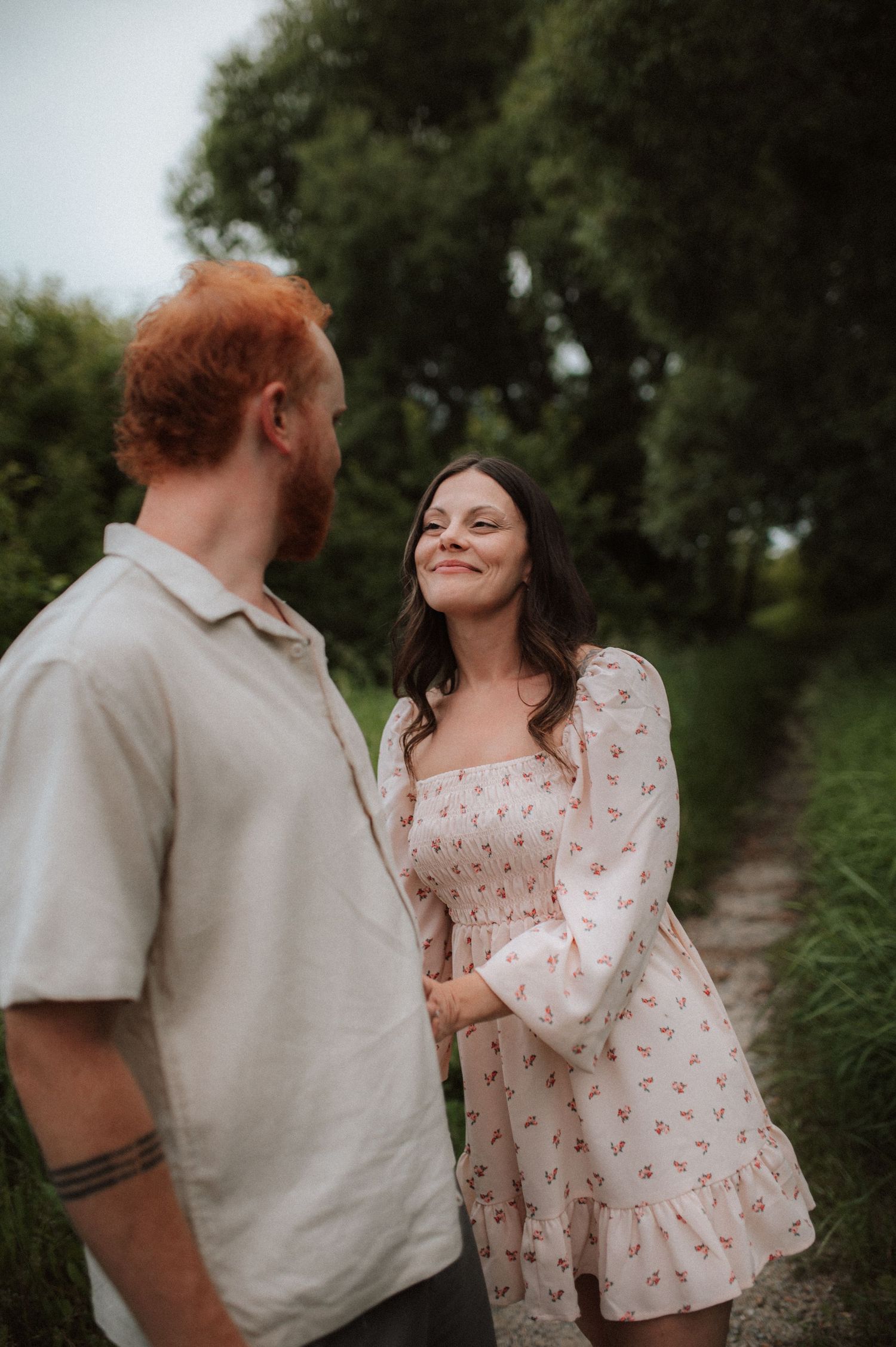 A couple shares a tender romantic moment outdoors on a path surrounded by greenery on a cloudy day.