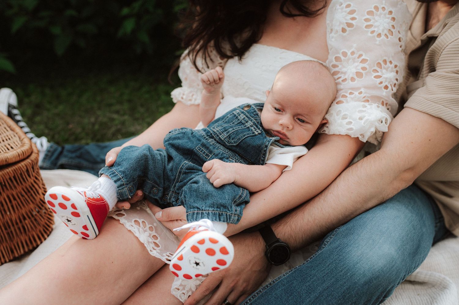 Baby resting on lap with picnic basket and heart-patterned items visible in outdoor garden setting.