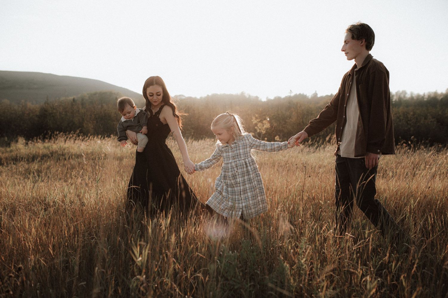 Family of four holding hands while walking through golden sunlit field at sunset in rustic countryside setting.