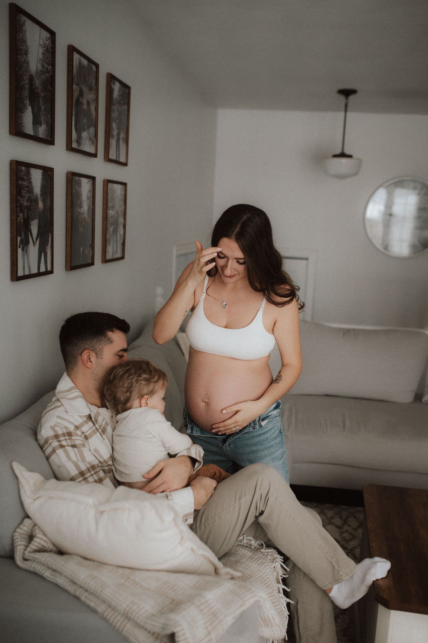 A tender family moment captured in a living room with natural light and framed wall art, featuring a pregnant person in white.