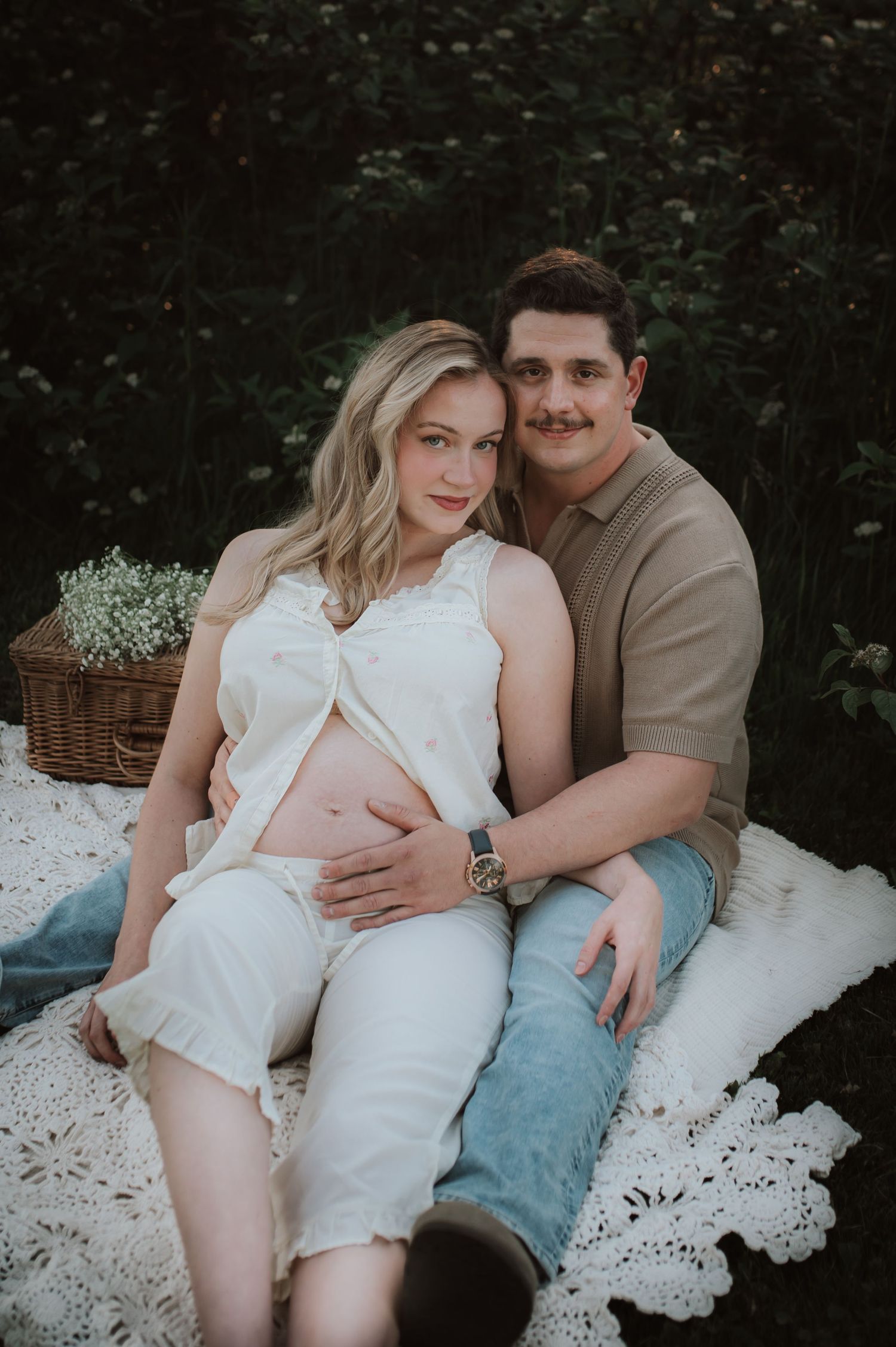 Expectant couple sits on white lace blanket during outdoor maternity photoshoot with flower basket in background.