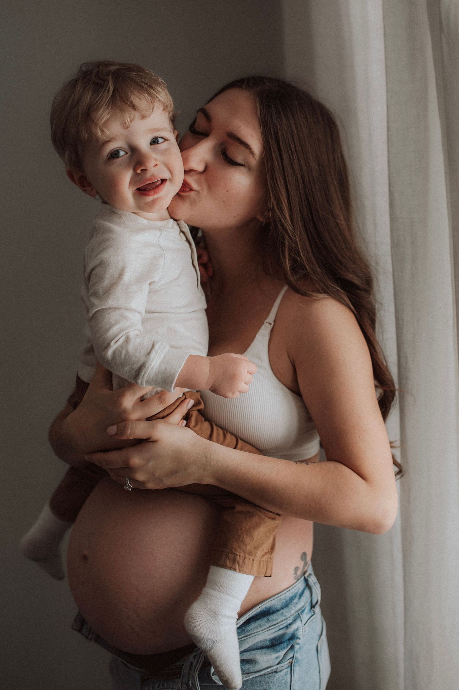 A person in denim holds and kisses a small child in white clothing against a neutral background.