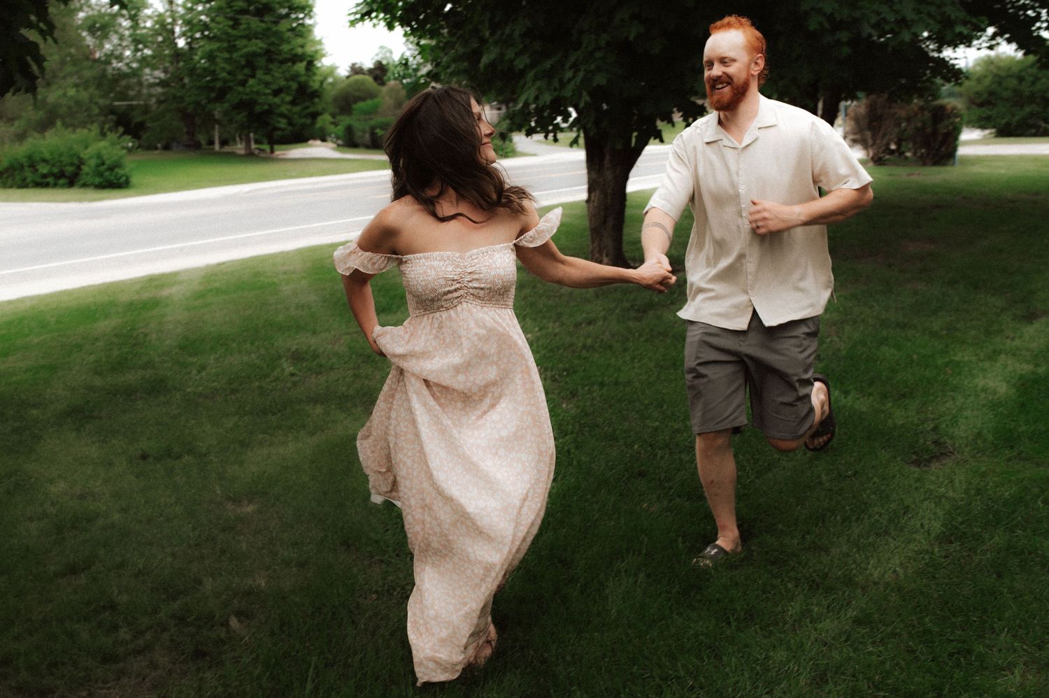 A person in a flowing blush dress runs playfully through a lush green garden while being chased.