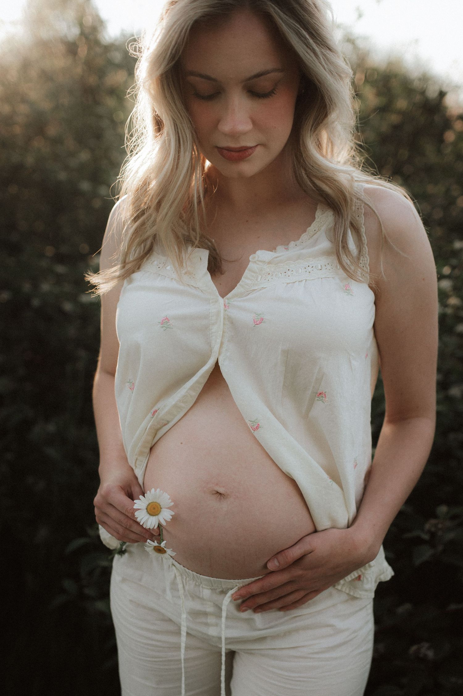 A serene maternity photo shows an expectant mother in white holding a daisy against her bare belly at sunset.