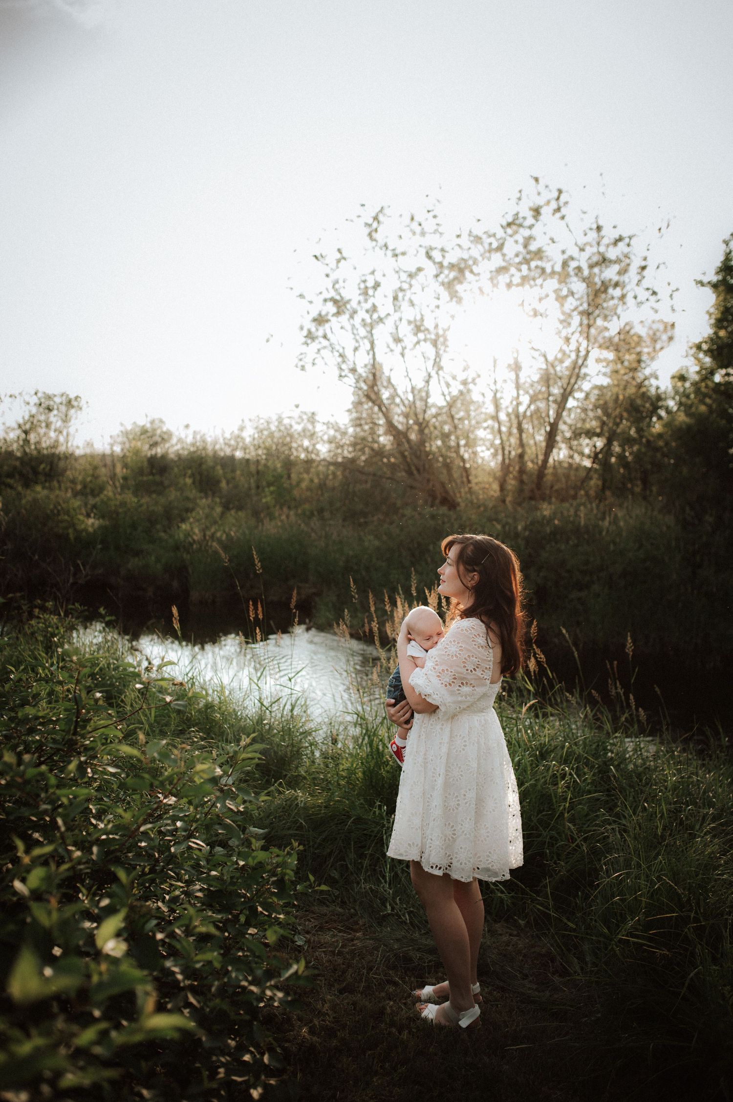 Silhouetted figures in white dresses stand by a serene lake at sunset amid tall grass and trees.