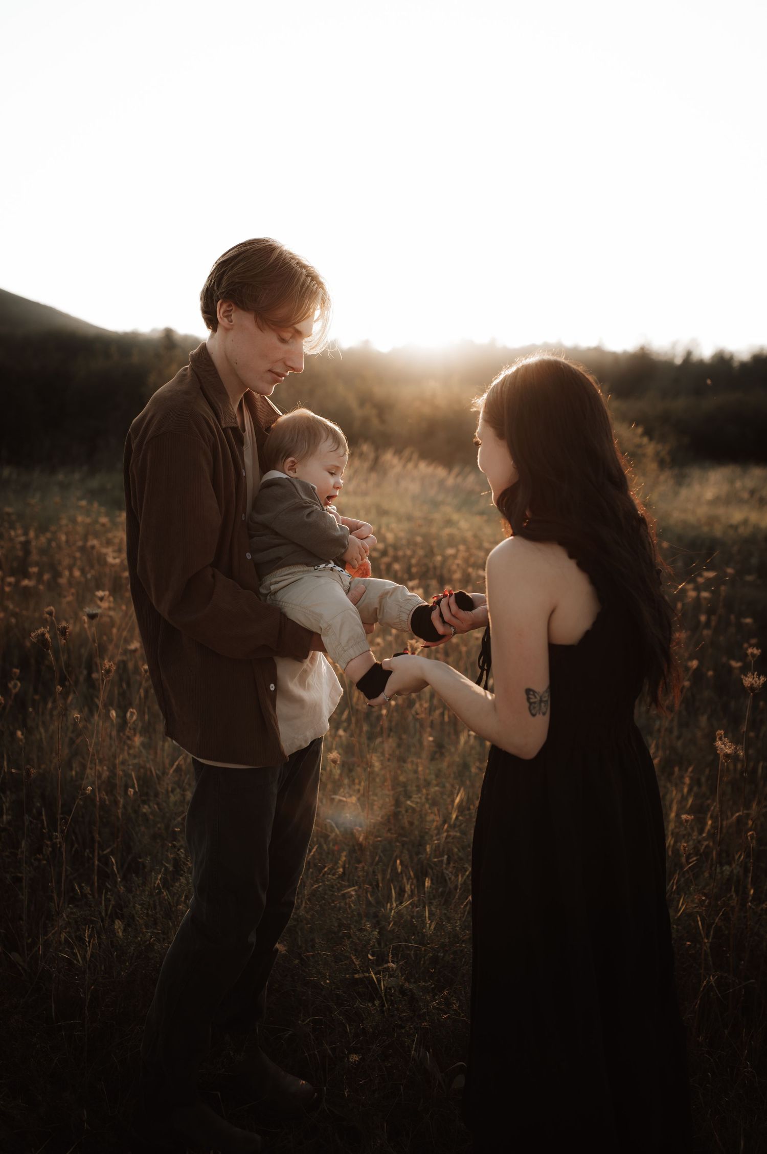 A couple shares a tender moment with their baby in a golden sunlit field at sunset.