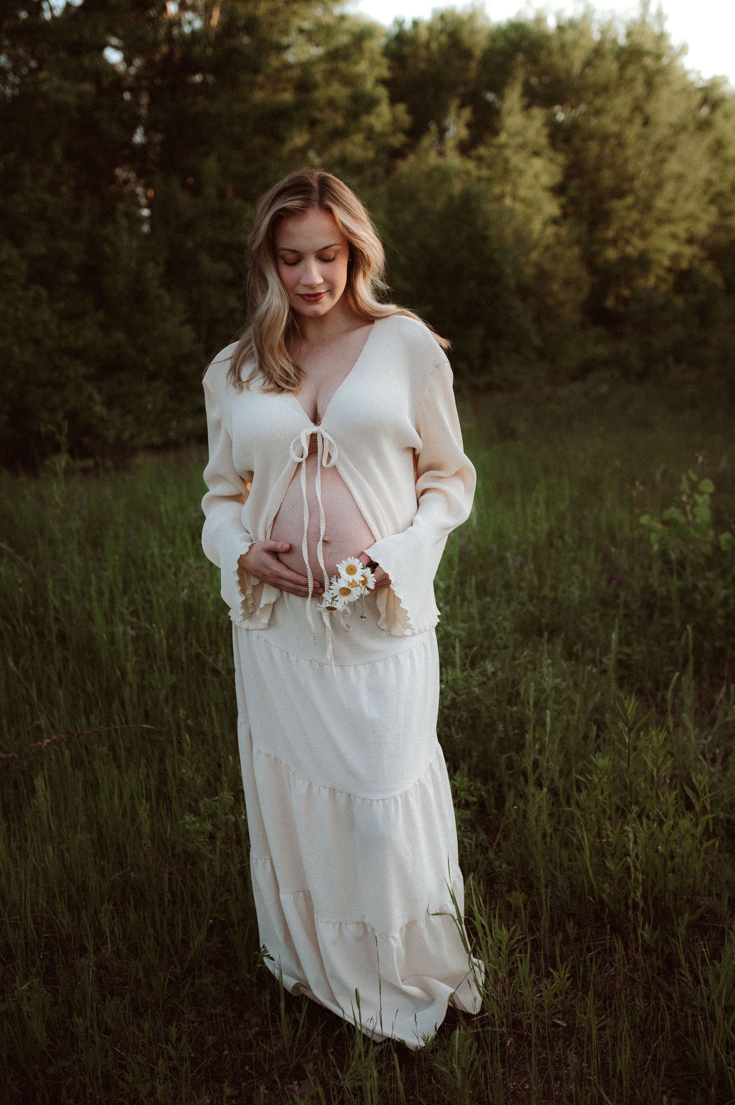 A woman in a flowing white dress and sweater stands in a field of tall grass at sunset holding wildflowers.