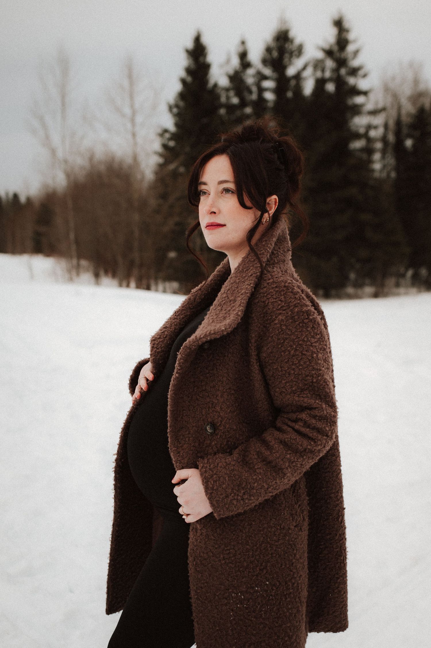 Person in a brown teddy coat poses against a snowy winter landscape with evergreen trees in the background.