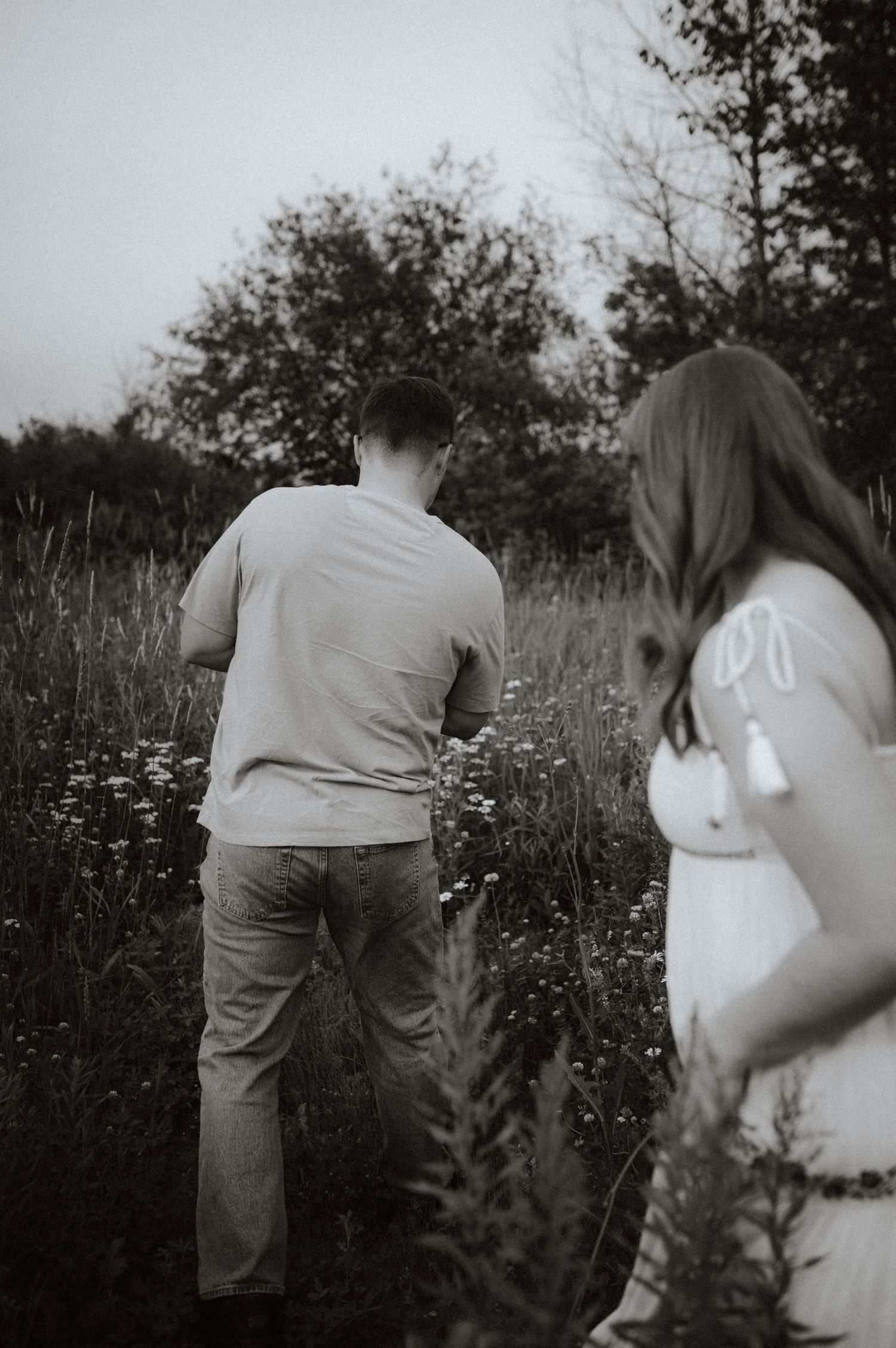 Black and white photo of couple standing in a wildflower meadow during golden hour with trees in background.