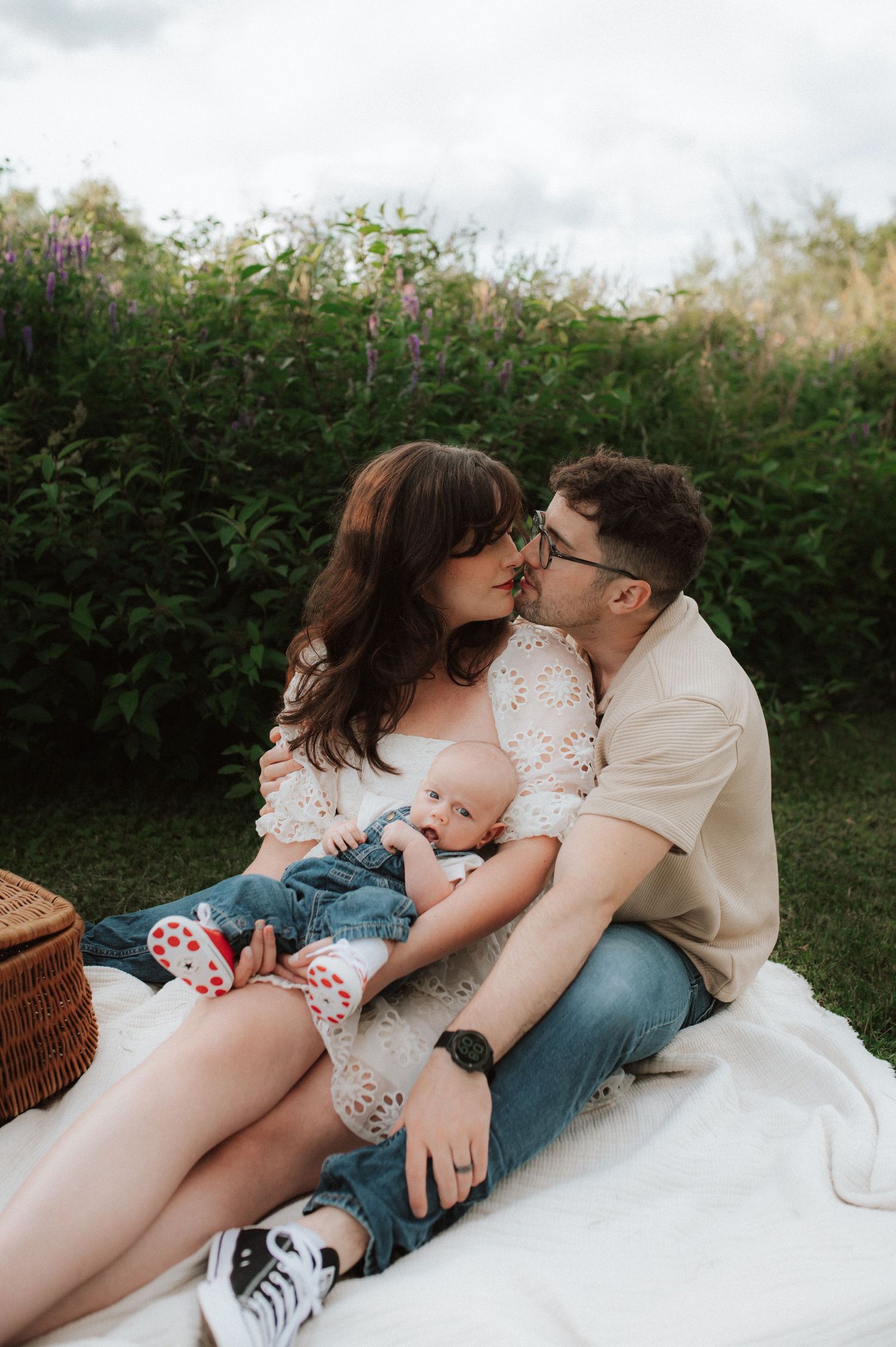 A couple shares an intimate moment on a picnic blanket with their baby in a garden setting surrounded by greenery.
