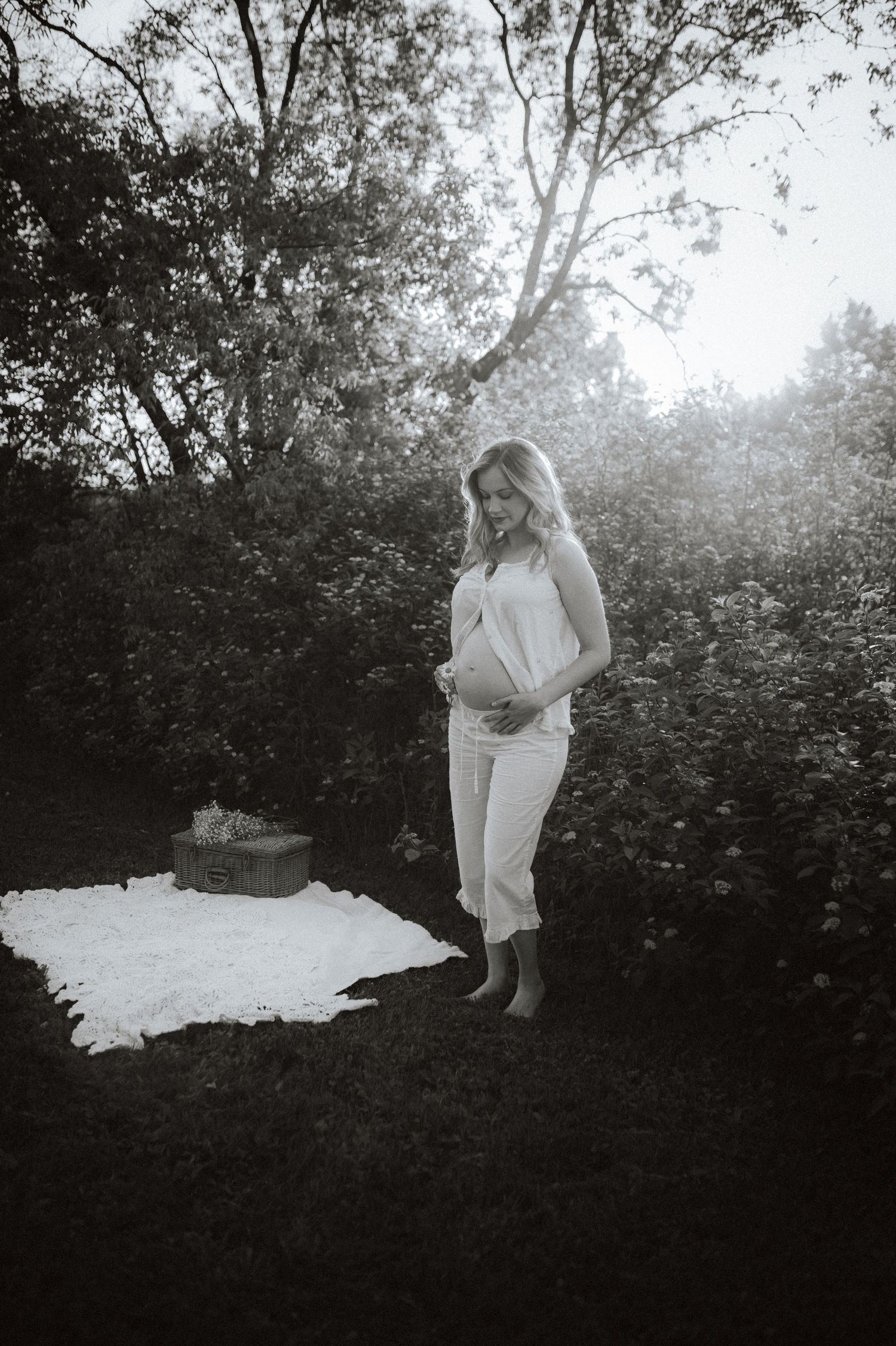 Black and white maternity photograph in a garden setting with a blanket on the ground and sunlight filtering through trees.