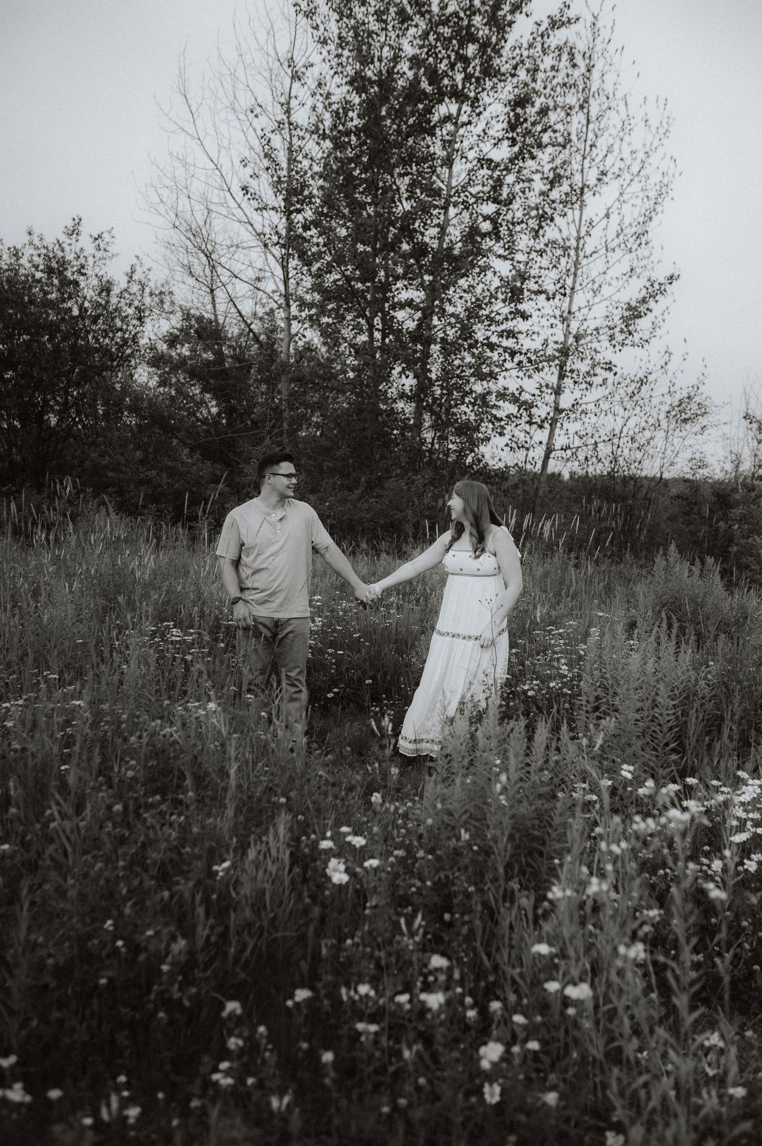 A black and white photo of a couple holding hands in a wildflower meadow with tall trees in the background.