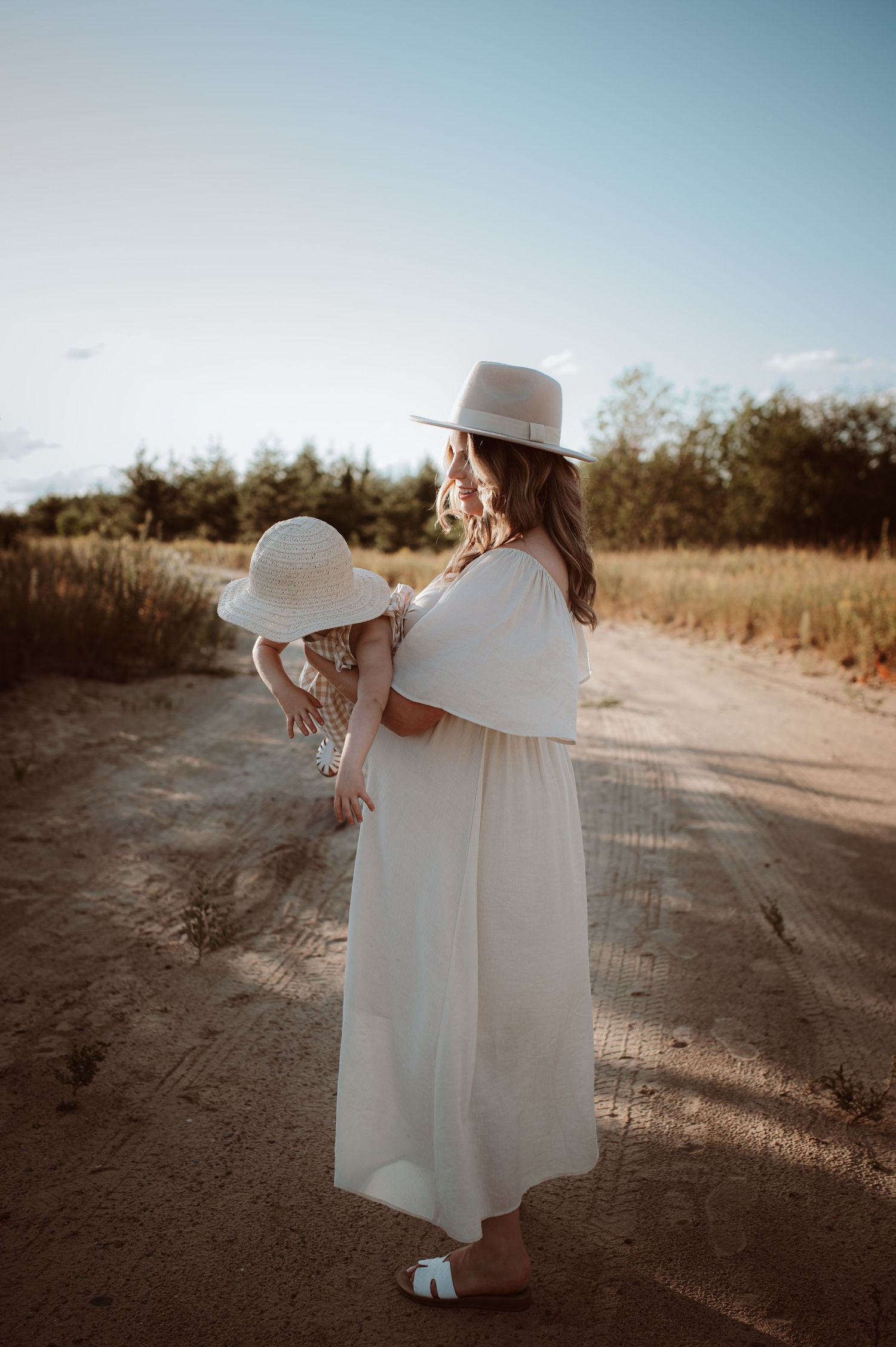 Two figures in flowing white dresses and sun hats walk hand in hand down a dusty dirt path at sunset.