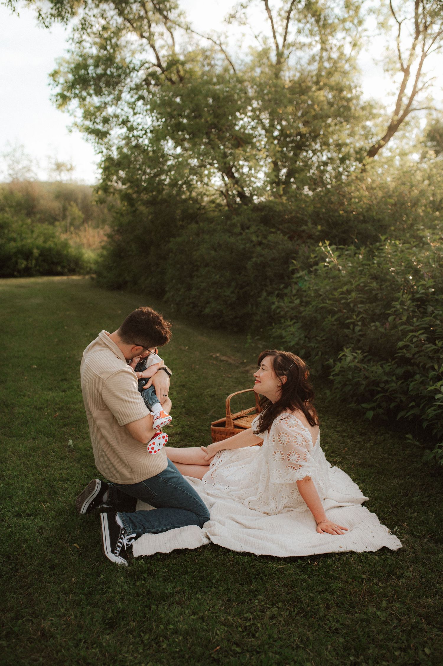 Two people sitting on a white blanket in a grassy outdoor area during golden hour with trees in the background.